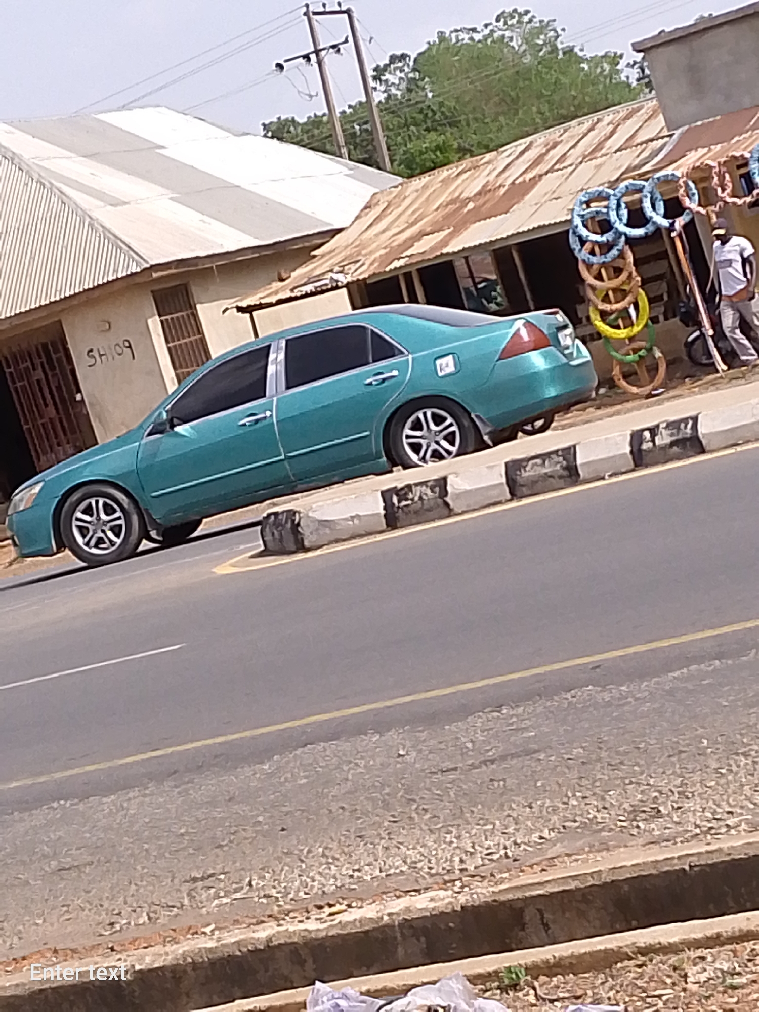 The image depicts a teal-colored sedan parked on the side of a road in an urban environment, possibly a small town or village. The vehicle is positioned adjacent to a curb marked with alternating black and white sections. Behind the car, there is a building with a corrugated metal roof, and next to it, a small stall displays colorful circular objects, possibly decorative items or toys, hanging along its storefront. A person dressed in a white shirt and dark pants stands near the stall, appearing to interact with it. The road is asphalt with a yellow line marking the edge. In the background, utility poles with power lines are visible, along with trees and more buildings. A sign with the letters "SH09" is on the building to the left of the car. The location is indicated as Unknown, Unknown.