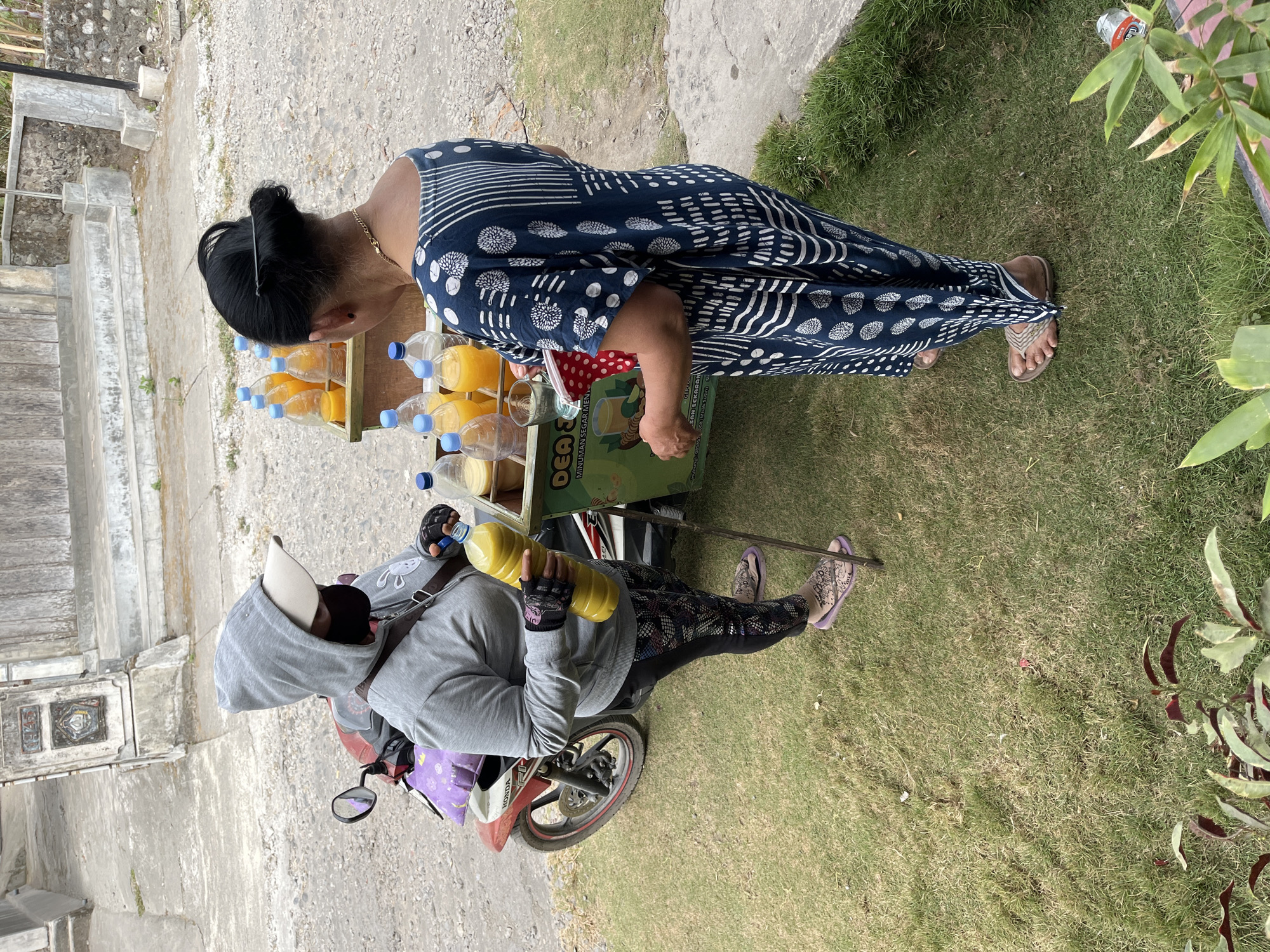 In Slorok, Indonesia, two women are seen near a cart filled with bottles of juice. One woman, wearing a grey hooded sweatshirt and a face mask, is on a red and white motorcycle. She is holding a large yellow bottle of juice. The second woman, wearing a dark blue dress with a white pattern, is standing next to the cart, holding a glass and a red polka-dot cloth. The cart is green and displays text, including "IDEA SEGAR" and "MINUMAN SEGAR MENTARI". The ground is covered with grass, and a stone pathway is visible in the background.