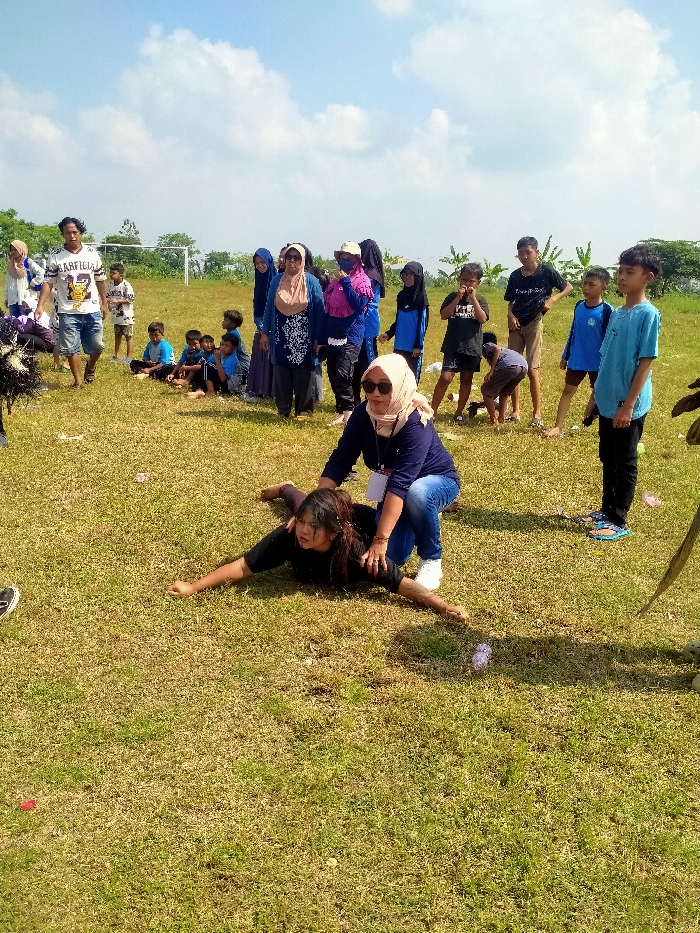 An outdoor scene in Celep, Indonesia, depicts a group of individuals gathered on a sunny, grassy field under a bright blue sky with scattered white clouds. In the foreground, a young woman with dark hair, wearing a dark long-sleeved shirt, lies prone on the grass with her arms outstretched to the sides. An adult woman, wearing a light-colored hijab, sunglasses, a dark long-sleeved top, blue jeans, and white sneakers, kneels over the prone woman, placing her hands on her back/shoulders, appearing to assist or guide her. In the mid-ground and background, a group of approximately 15-20 adults and children observe the activity. This group includes individuals in various casual attire; some women wear hijabs, while others wear t-shirts, shorts, and jeans. Some children are seated on the grass, while others stand watching. Notable figures in the background include an adult male in a white "GARFIELD 22" t-shirt and blue shorts walking to the left, and a young boy in a light blue t-shirt standing on the right. A white soccer goalpost is visible in the far-left background, beyond which are trees and foliage. Small pieces of litter are scattered across the grass.