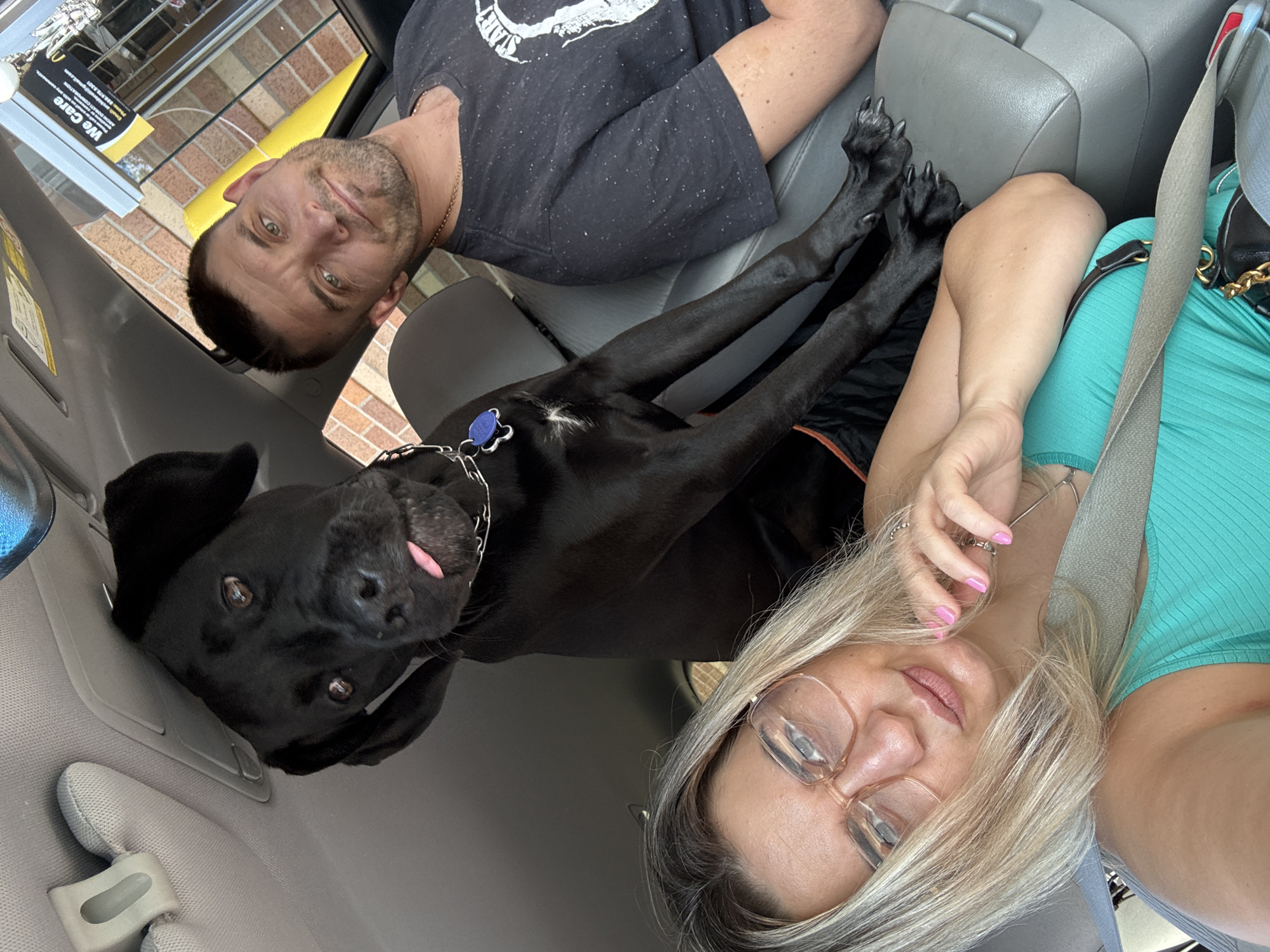 The image shows three subjects in a vehicle, possibly at a drive-through window for a fast-food establishment such as McDonald's, suggested by the "We Care" sign visible in the background which is a McDonald's initiative. A black dog with its tongue sticking out is positioned between a woman and a man. The woman, in the foreground, wears glasses and a turquoise tank top, with her blonde hair falling over her face. She is resting her hand on her chin, with pink painted fingernails. The man, in the background, has short brown hair and a beard, wearing a dark t-shirt with a graphic design. The dog's paws are resting on the center console of the car. The interior of the car is visible, including the car's ceiling and sun visor. The background outside the car suggests a brick building and the drive-through structure.