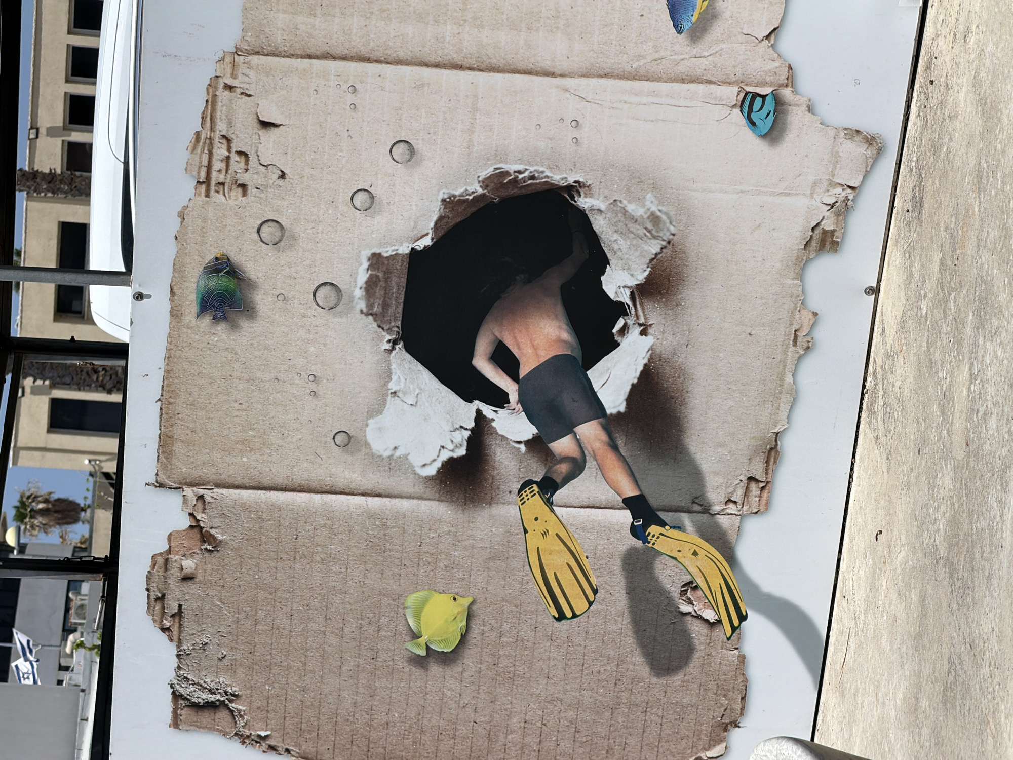 The image depicts a collage on a wall, featuring a ripped cardboard cutout with a person diving through the hole. The person is wearing black shorts, socks, and yellow flippers. Several fish are pasted around the cutout, including a yellow fish near the bottom, a blue fish at the top right, and a striped fish on the left. Bubble shapes are also pasted around the hole. The wall is white with a textured gray surface visible on the right. The backdrop includes a building reflected in glass, with an Israeli flag visible on the left. The location is Tel-Aviv, Israel.