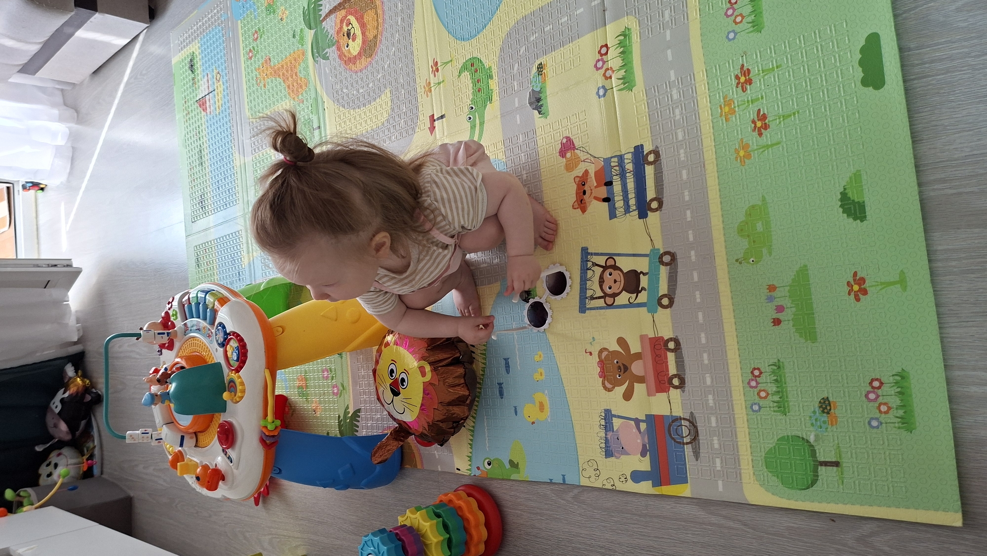 A toddler with light brown hair tied in a small topknot is kneeling on a colorful play mat. She wears a striped shirt and light pink shorts. She is interacting with a pair of white sunglasses on the mat. Around her are various toys including a large activity center with colorful features, a deflated lion-shaped balloon, and a rainbow stacking toy. The play mat is segmented with different animal themes and street patterns. The background shows a window with natural light and a glimpse of a white surface or object. Based on the prompt, the location is Chaikovsky, Russia.