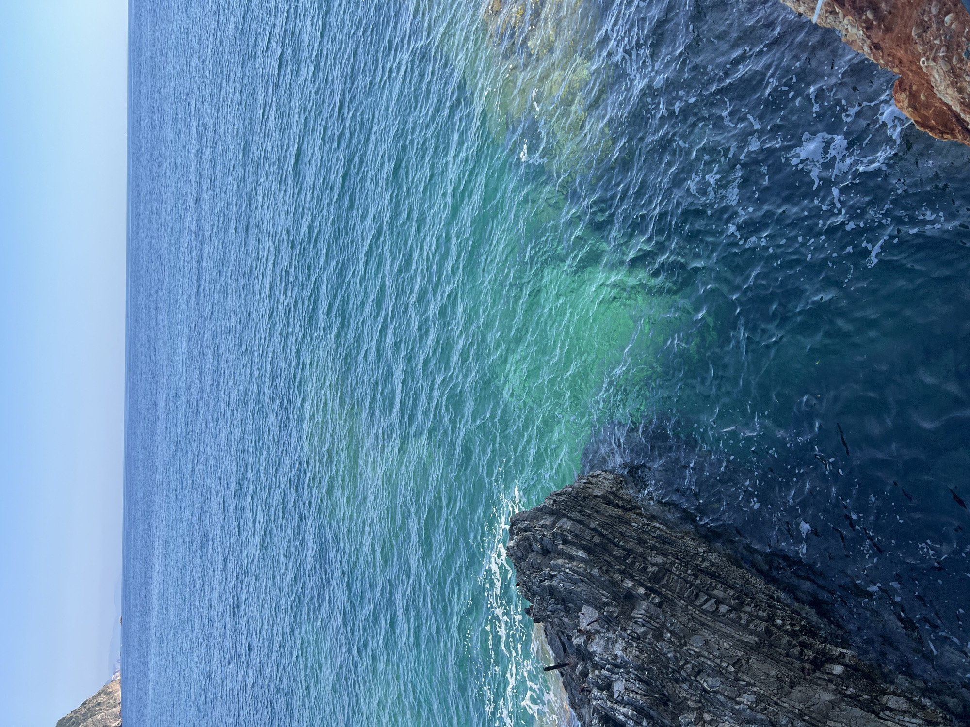The image shows the Adriatic Sea meeting the rocky coastline near Budva, Montenegro. The sea transitions from a pale blue in the distance to a vibrant turquoise near the shore, and finally to a darker blue where it meets the rock. A rock formation with clear stratification is located on the lower right and in the middle. Another rock formation is visible in the upper right of the frame, with a reddish-brown hue. The horizon is mostly flat, with the light blue sky peeking through on the far left.