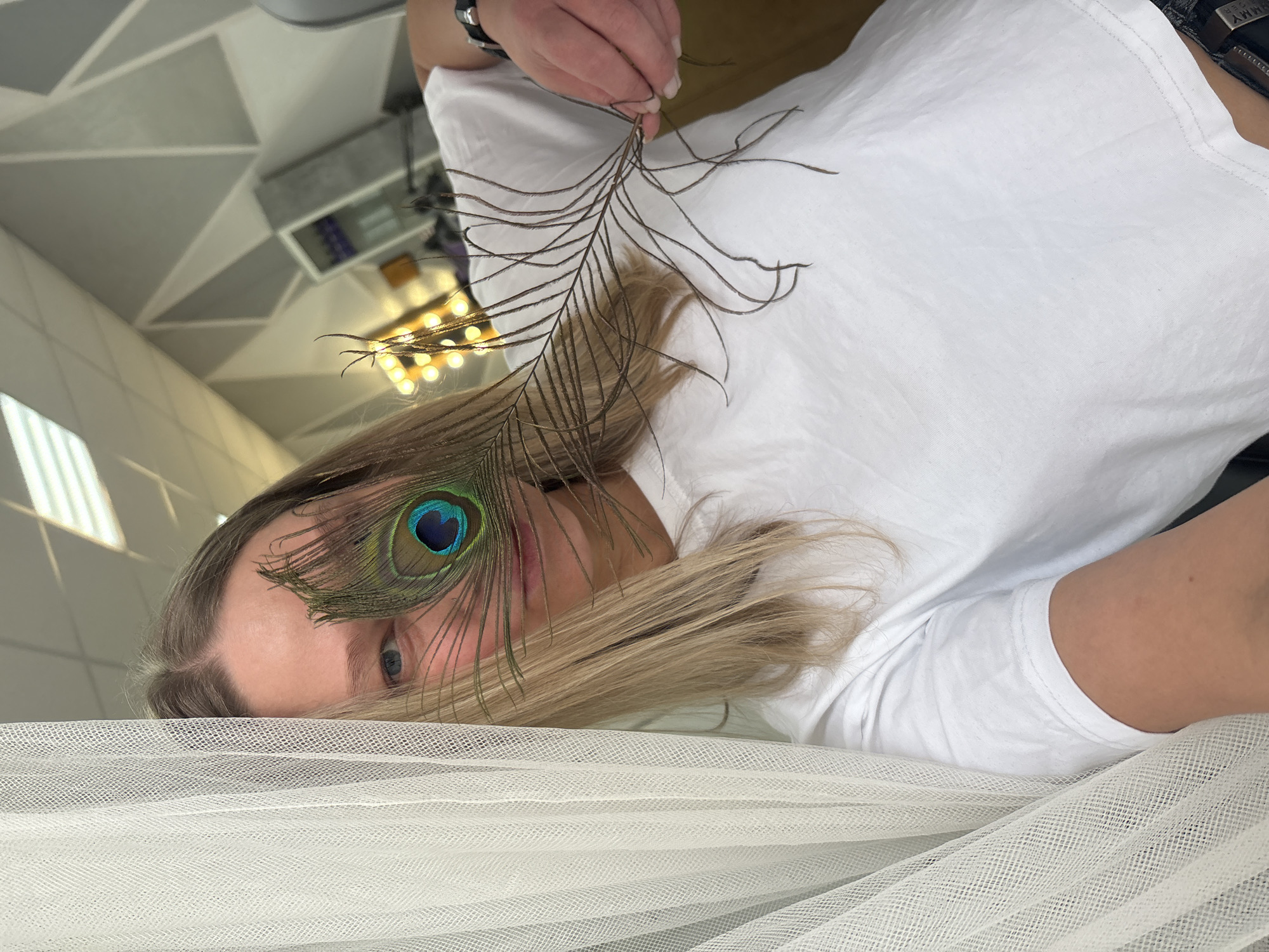 A woman lies on a white, semi-transparent, net-like fabric. She is wearing a white t-shirt. A peacock feather is positioned near her face, partially covering one eye. Her hair is blonde. She is holding the upper part of the feather in her hand. The background shows an interior with lighting fixtures and geometric ceiling patterns. Based on the visual clues, the location is likely indoors, potentially a studio or event space, but not explicitly identified as Perm, Russia.