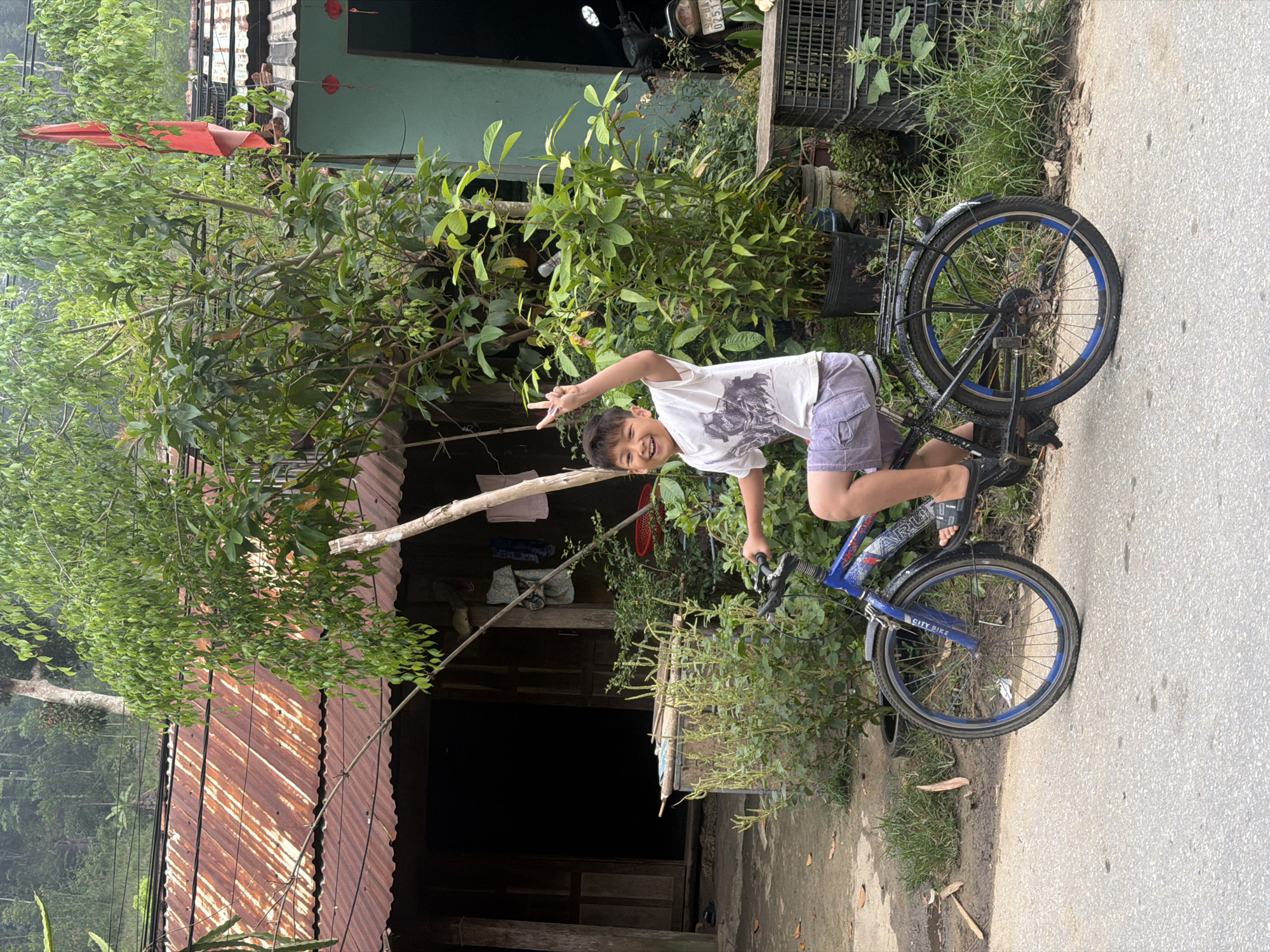 A young boy, likely around 8-10 years old, is posed on a blue bicycle, gesturing with a peace sign and smiling. He is wearing a white t-shirt and grey shorts. The bike is a "City Bike" with blue rims. The setting is a rural area with a weathered building, likely a home, made of wood and corrugated metal. There is lush vegetation, including trees and shrubs, surrounding the building. A concrete or paved road is visible to the right of the frame. A red flag hangs from the building, possibly indicating a local custom. The environment suggests a location in a rural or semi-rural area of Vietnam, but it is not possible to verify if it is the city of Da Nang. A partially obscured motorcycle is parked near the building.
