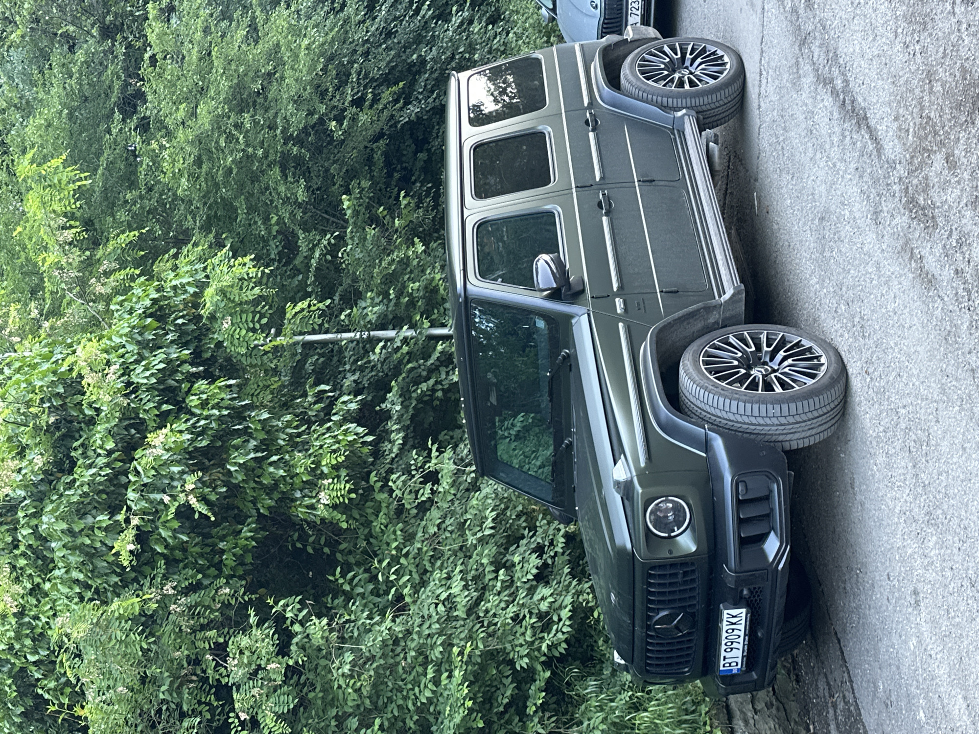 A dark green Mercedes-Benz G-Class SUV is parked on a narrow paved road. The vehicle is parked close to a lush green area with dense trees and shrubs. Part of another car is visible behind the SUV. The license plate of the SUV reads "BT 9909 KK", which indicates the vehicle is registered in Veliko Tarnovo, Bulgaria. The location appears to be a residential street with abundant greenery.