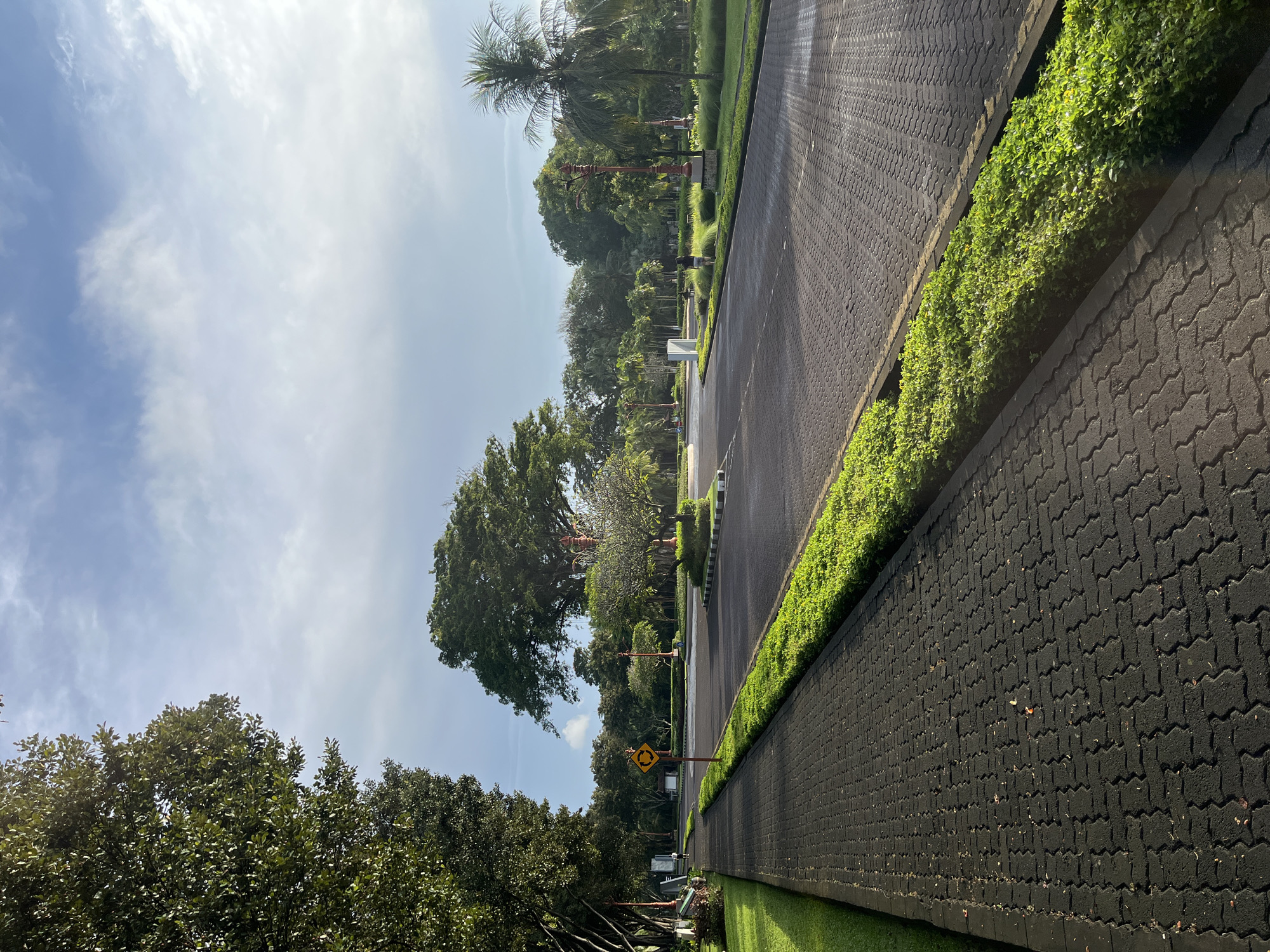 This media file captures an outdoor scene featuring a paved road or driveway flanked by meticulously maintained landscaping under a bright sky. The perspective is slightly angled, looking down the length of the road.
On the immediate right of the road, there is a narrow strip of neatly trimmed green grass, followed by a dark grey pathway constructed from interlocking pavers. Beyond this path, a raised planter box filled with dense, vibrant green bushes runs parallel to the road, backed by a tall wall also made of the same dark grey interlocking pavers.
To the left of the road, a wider expanse of green grass leads to a variety of lush tropical trees, including tall palm trees and other dense foliage, creating a verdant border. Several decorative lampposts or similar ornamental structures are spaced along this side of the road. In the middle distance on the left, a yellow diamond-shaped road sign with a black border and a black circular arrow icon indicates the presence of a roundabout or traffic circle ahead.
The sky above is a clear, bright blue with scattered, wispy white clouds, suggesting pleasant weather and good visibility. The scene appears serene and well-ordered, characteristic of a park, resort, or well-maintained institutional grounds. No people or activities are visible, emphasizing the tranquil setting. The time of day appears to be daytime, likely morning or early afternoon, with ample natural light. The tropical vegetation is consistent with the given location context of Kuta Selatan, Indonesia.