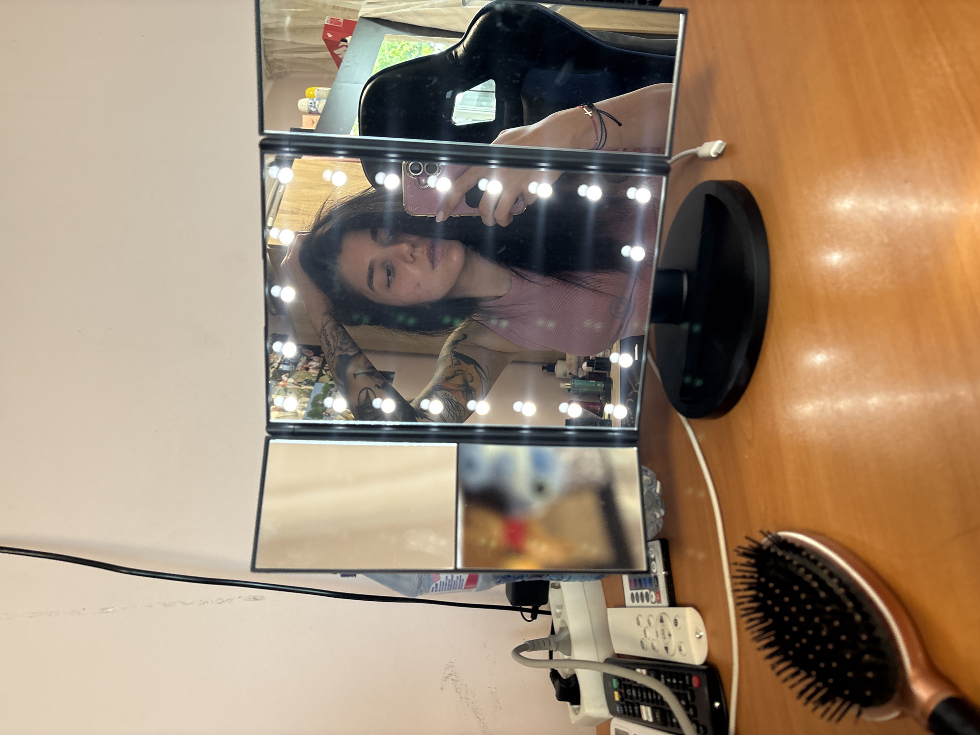 The image shows a woman taking a selfie in a lighted makeup mirror. The mirror is on a wooden surface, possibly a desk or vanity. A hairbrush, remote controls, and an electrical outlet strip are also on the surface near the mirror. The mirror's reflection shows parts of a room, including a black chair and various items on a shelf. The location of the image is not possible to determine.