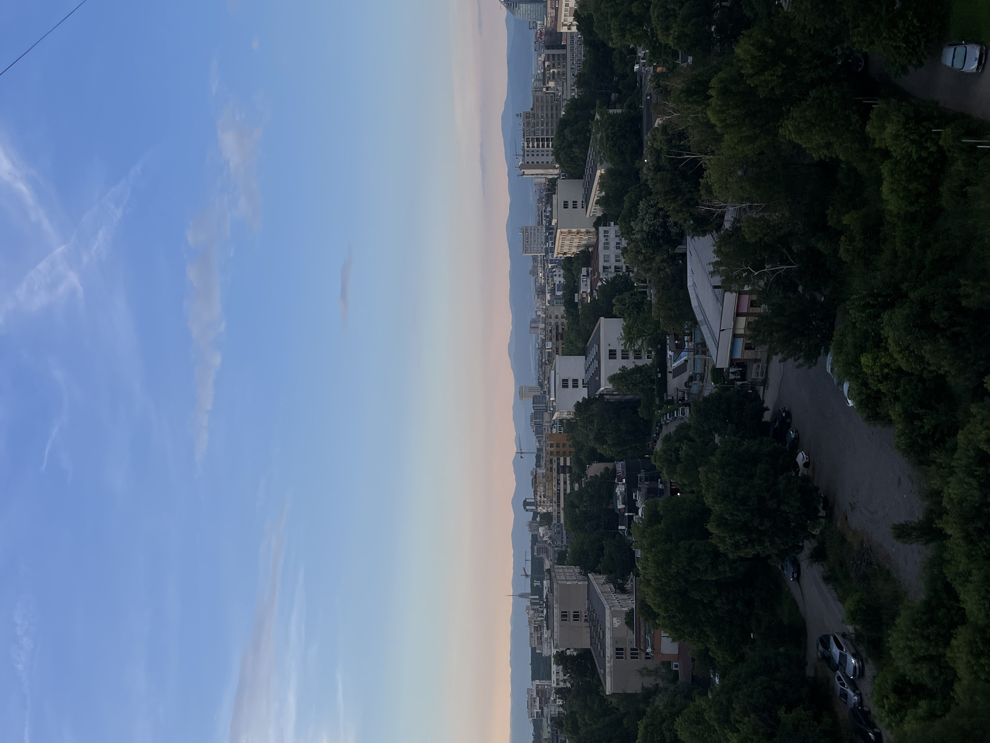 This is a high-angle, full shot of a city skyline at dusk. The sky is a gradient of blue and light pink. In the foreground, lush green trees are densely packed, obscuring the lower portions of buildings. The middle ground reveals a mix of apartment buildings and other structures, some appearing older and some more modern, with varying architectural styles. Beyond these, a more distant cityscape stretches out, characterized by taller buildings, and a prominent tower with a spire. In the far distance, a mountain range is visible against the horizon. There are several cars parked and driving on roads visible between the trees and buildings. The scene is quiet and appears to be a residential or mixed-use area within a city, likely during the evening hours due to the lighting.