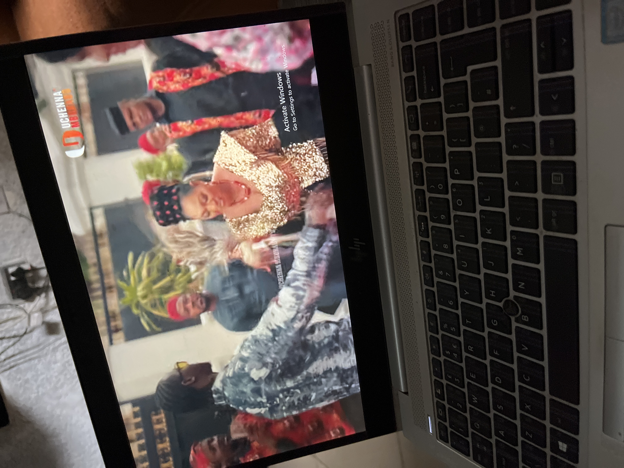 An angled shot displays a silver laptop with a black keyboard, open and showing a video on its screen. The laptop features "BANG & OLUFSEN" branding above the keyboard. The screen prominently displays a "Activate Windows Go to Settings to activate Windows" message.
The video being watched features several people, likely portraying a scene from a movie, music video, or cultural event, consistent with the Abuja, Nigeria context. Visible figures include a woman in a shimmering gold, sequined top and a red necklace, looking down. Another person wears a black garment with red and gold patterns and a red cap. Other individuals in traditional or festive attire, including one in a camouflage-patterned top, are also seen. A logo "UCHENNA MBURANA" is visible in the upper left corner of the video playback.
The image captures the laptop in an indoor setting, with soft, ambient lighting. The keyboard shows standard QWERTY keys with numbers and symbols.