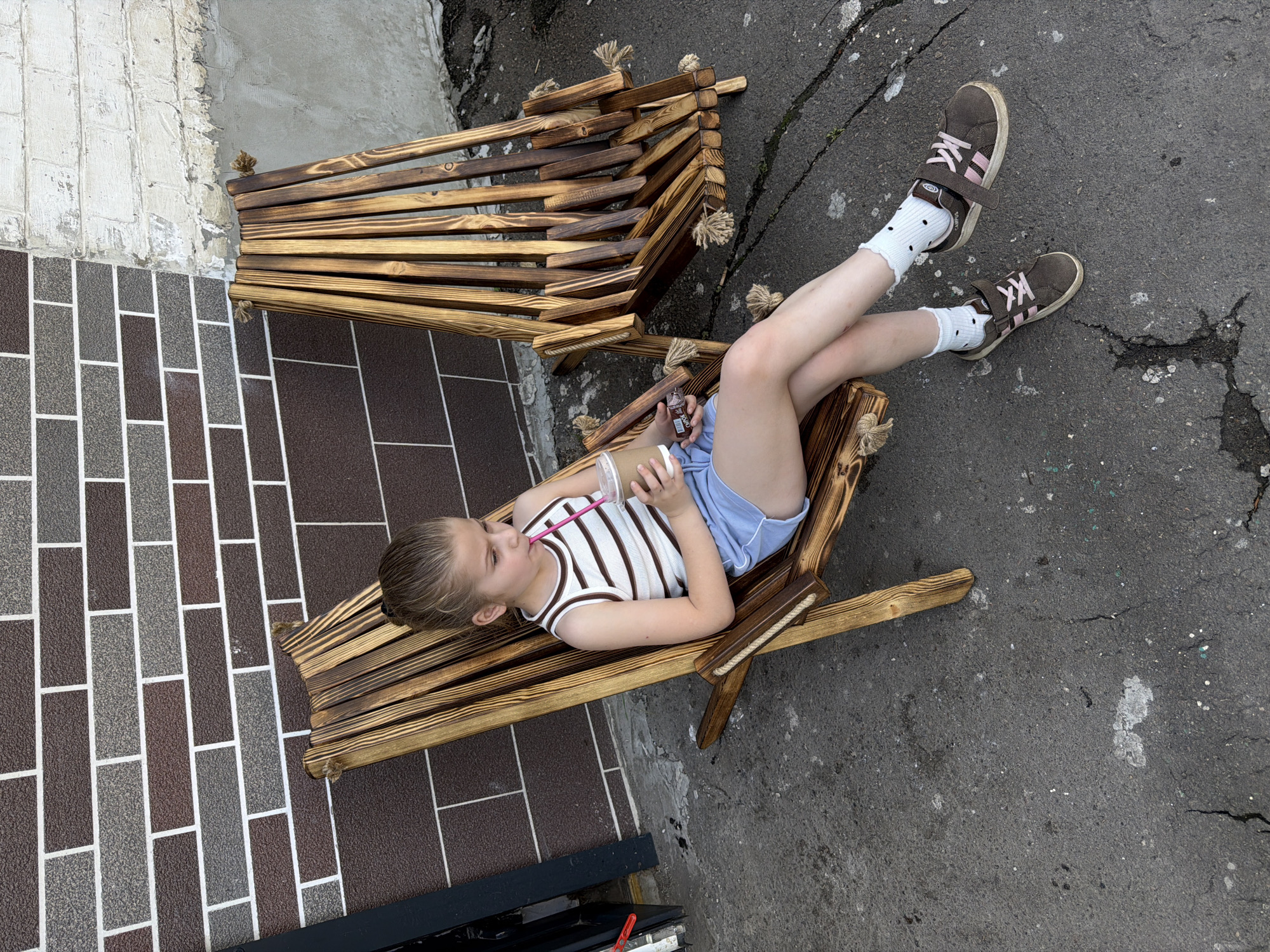A young girl with blonde hair pulled back in a ponytail sits in a wooden lounge chair, holding a cup with a straw and a chocolate bar. She is wearing a striped tank top, light blue shorts, white socks with black polka dots, and brown sneakers with pink laces. She is facing slightly to her right, looking down at the cup. Behind her, another identical wooden lounge chair is empty. The chairs are positioned on a cracked asphalt surface in front of a brick wall with two different colored brick patterns. To the right of the brick wall is a section of rough, white-plastered brickwork. The overall environment suggests an outdoor seating area, possibly at a cafe or restaurant.