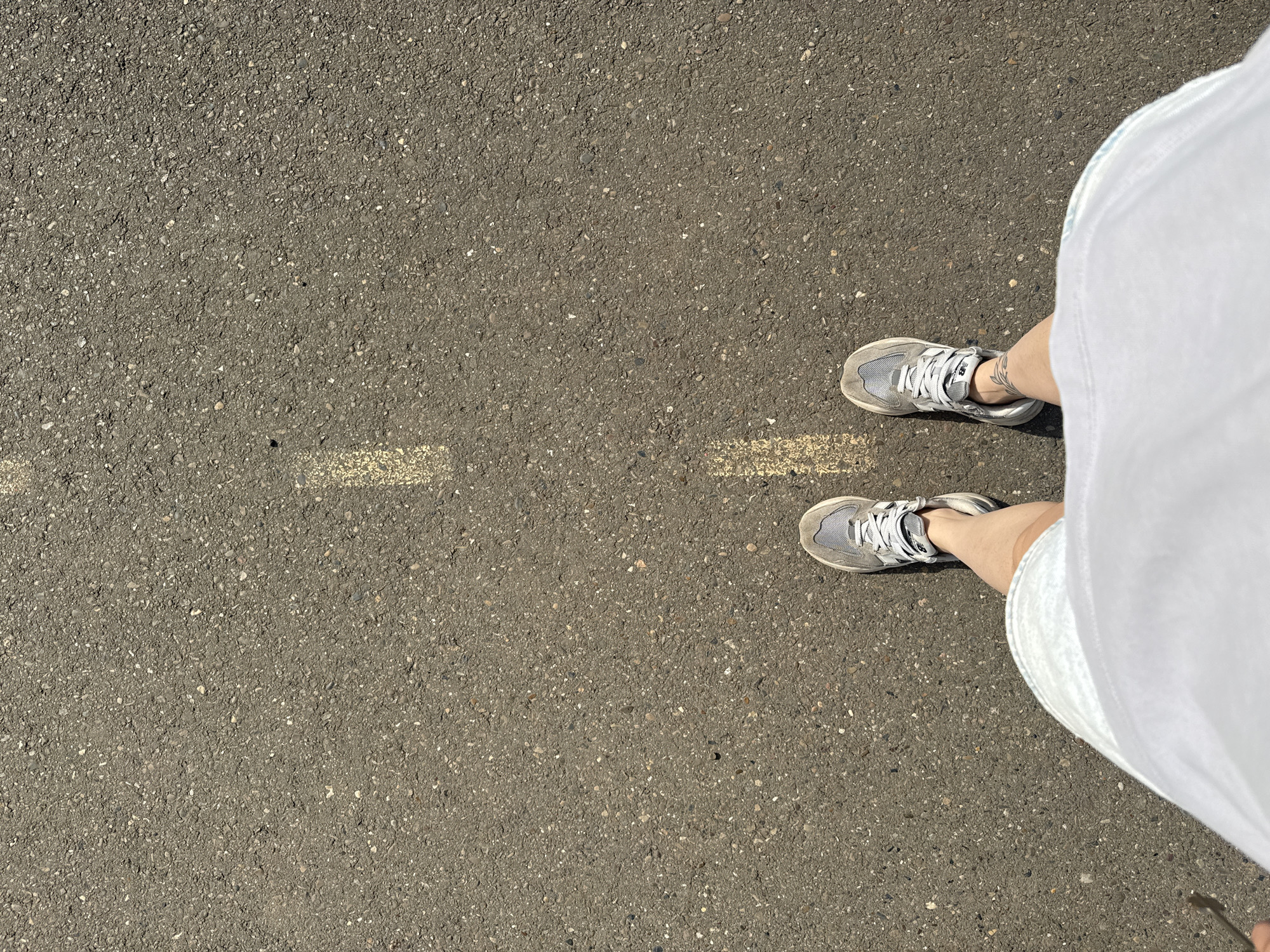 The image shows a view from above of a person standing on asphalt. The asphalt is grey and has a broken yellow line on the left side of the frame. The person is wearing grey sneakers and a white dress. The location cannot be verified as Chaikovsky, Russia based solely on this image.
