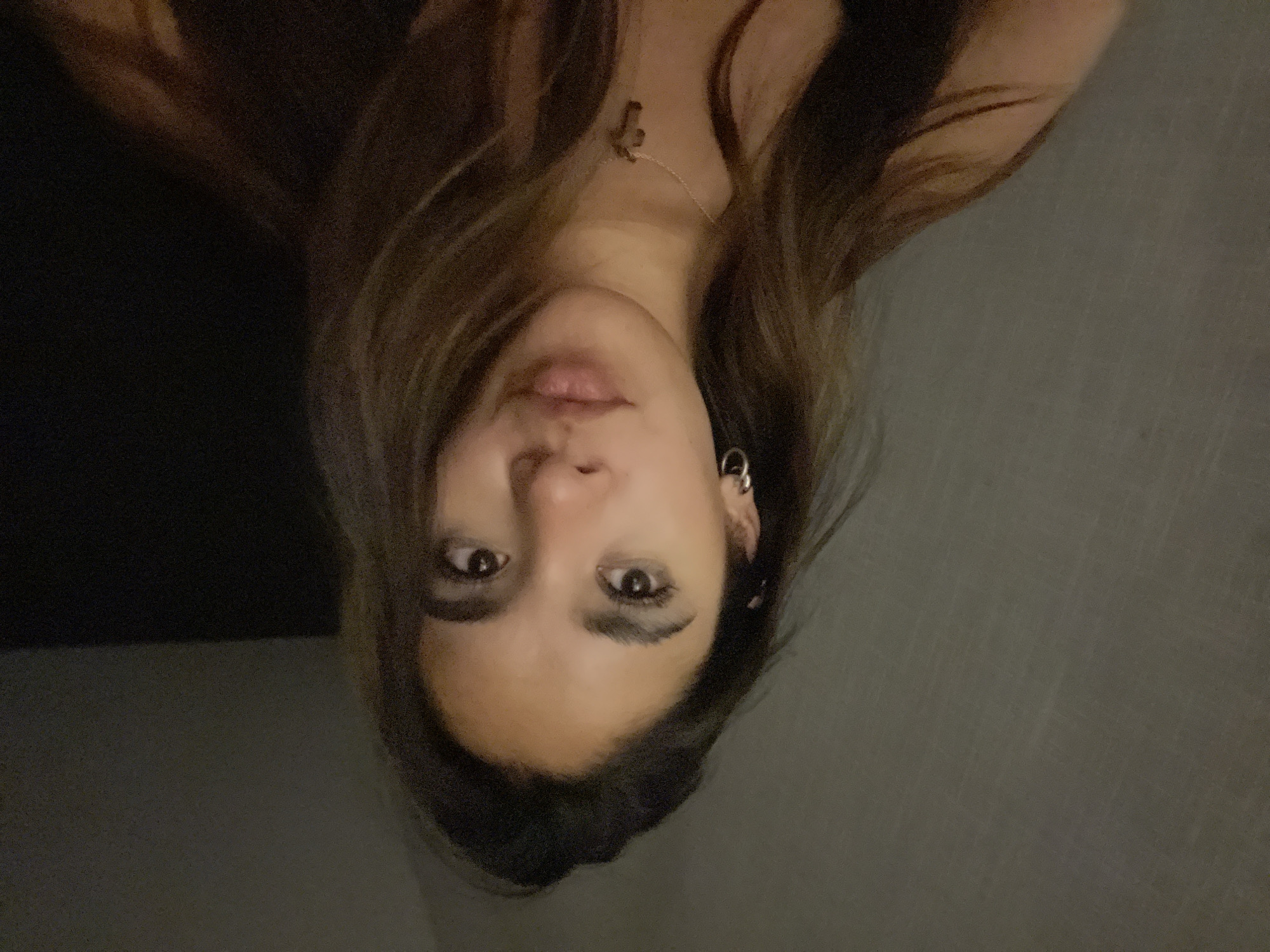 A person is visible from the chest up in an indoor setting. The person has long brown hair, brown eyes, and fair skin. They are wearing a necklace with a pendant and an earring on their left ear. The background appears to be a dark wall. The image is dark and slightly blurred. It is upside down. There is no information in the image to suggest the location is San Bernardino Tlaxcalancingo, Mexico.