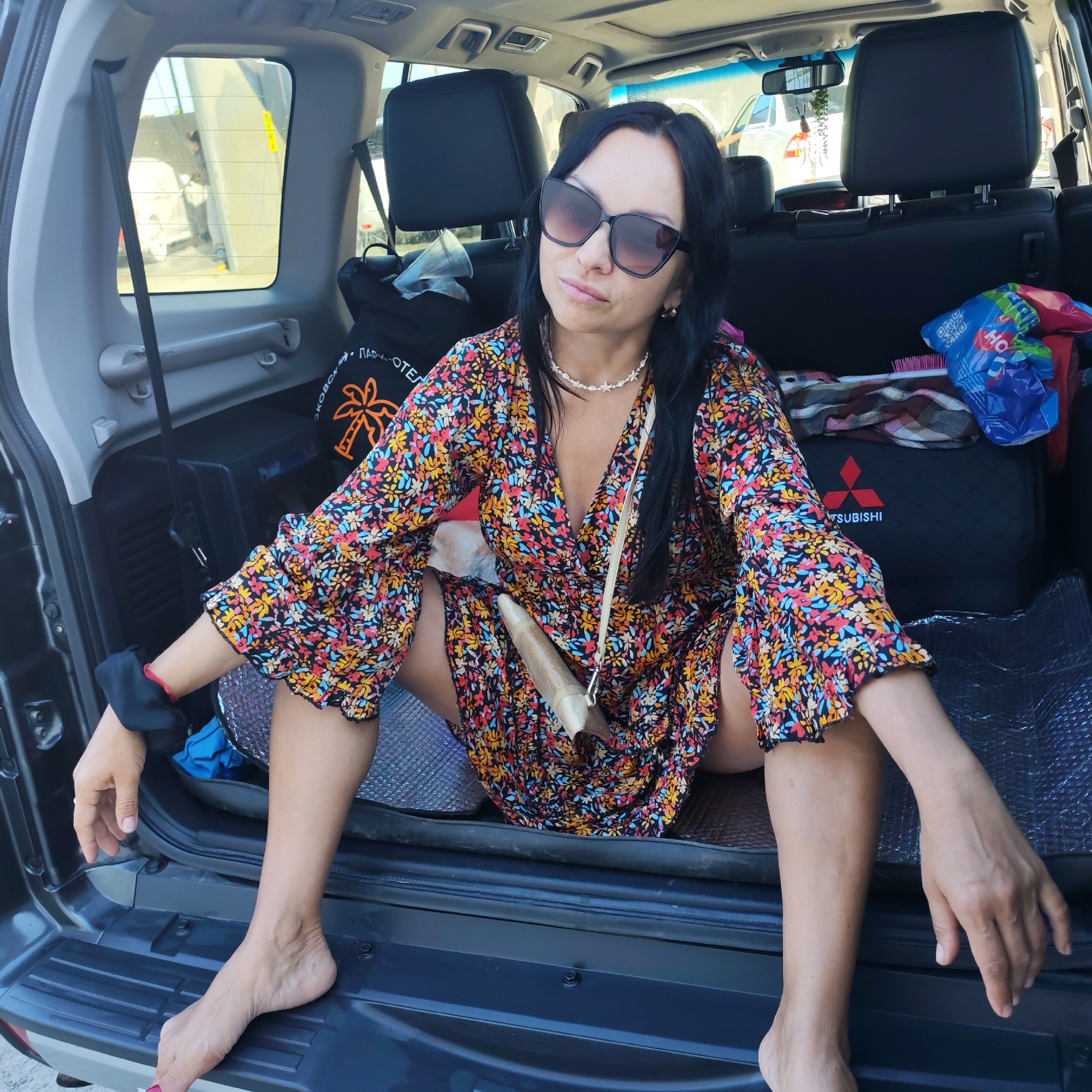 The image shows a woman with dark hair and sunglasses sitting in the open trunk of a dark-colored SUV. She is wearing a colorful floral dress and a light-colored beaded necklace. A small beige purse is slung across her body. Her left leg is bent, and her feet are visible. Behind her, the interior of the SUV is visible, including car seats, bags, and a pink hairbrush. Outside the vehicle, through the open trunk, a bright, possibly sunny environment can be seen, with what appears to be a white car in the background and a building structure. The scene suggests a relaxed, casual setting, possibly a stop during a trip or an outing. There is no discernible information in the image that would place this scene specifically in the city of Pushkino, Ukraine.