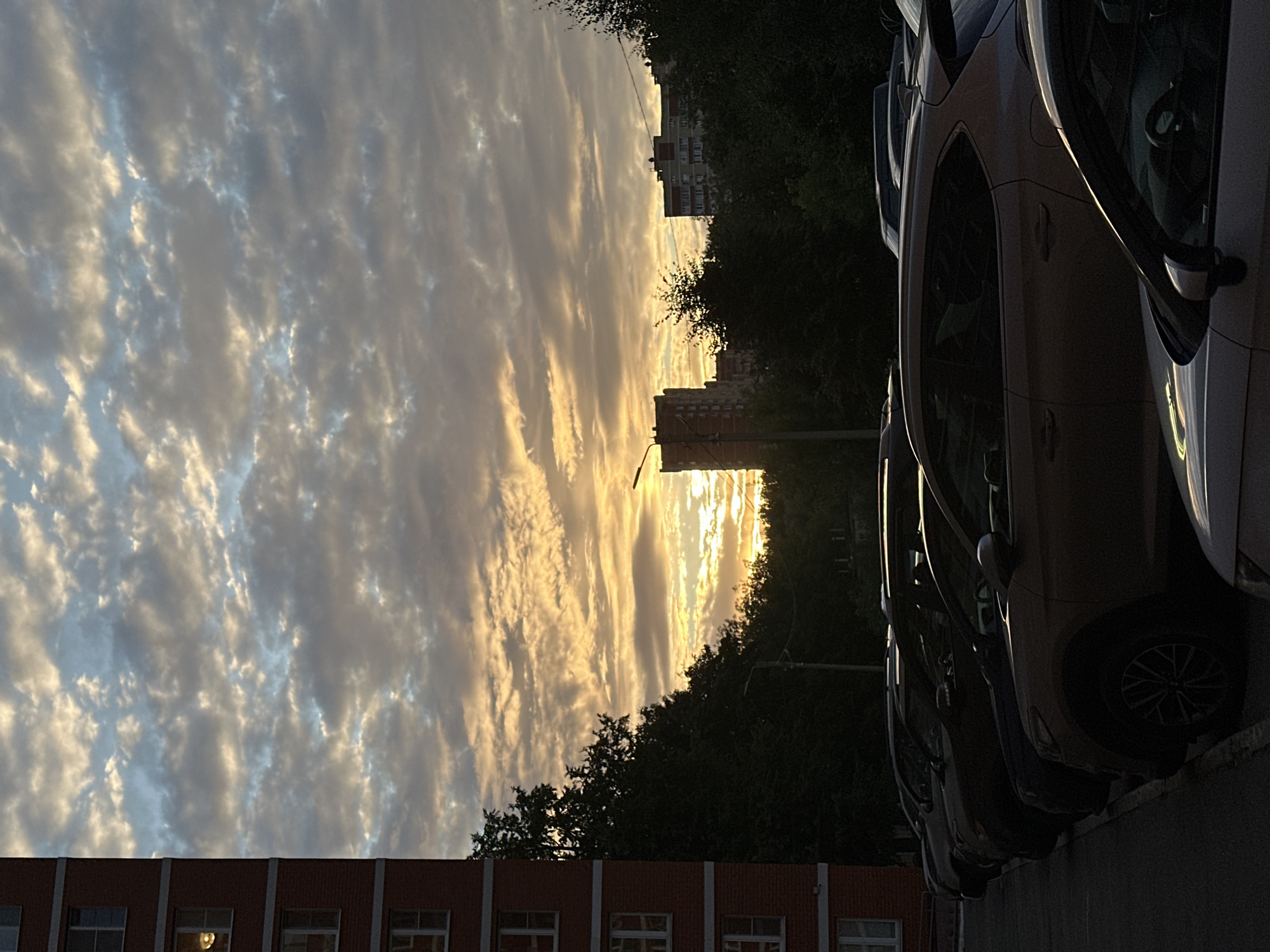 This is a medium-format, vertical, eye-level shot taken at sunset in the city of Shchyolkovo, Russia. The scene is dominated by a dramatic sky filled with textured clouds, illuminated by the setting sun, creating a warm, orange glow. On the left, the side of a tall, red brick apartment building with visible windows is cropped into the frame. In the foreground and mid-ground, a row of parked cars is visible, mostly silhouetted against the brighter background. The cars appear to be in a residential parking lot. Behind the cars, a line of dark green trees partially obscures the view of more distant apartment buildings and a cell tower. The overall atmosphere is one of an urban evening transitioning into night, with a focus on the natural beauty of the sky.