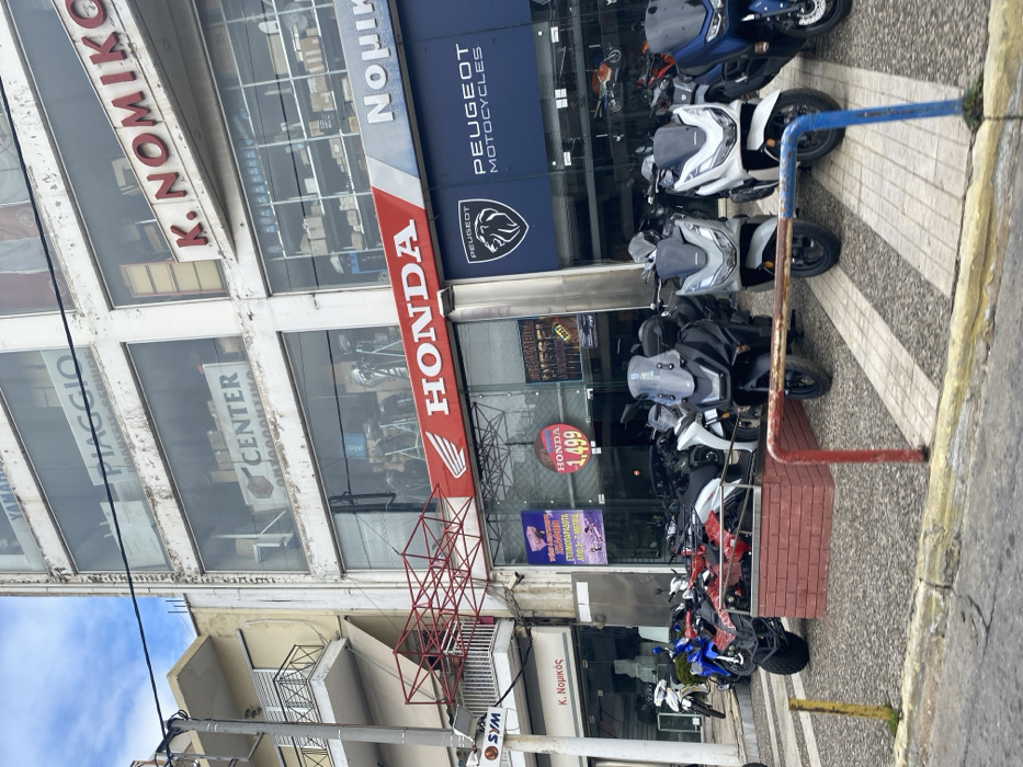 This daylight image captures the street view of a multi-brand motorcycle and scooter dealership, "K. NOMIKO," in Nikea, Greece. The building features large display windows on its lower floors, showcasing numerous vehicles.
Prominent signage includes a large, vertical red sign for "HONDA" with its logo, and a blue sign for "PEUGEOT MOTOCYCLES" with its lion emblem. Other visible brands indicate that the dealership also handles "PIAGGIO," "YAMAHA," and "SYM." The name "K. NOMIKO" is visible in multiple locations on the building.
A variety of motorcycles and scooters are displayed both inside the showroom and lined up on the sidewalk outside. These include multiple white and grey scooters, as well as different models from Honda and Peugeot. A special offer for a Honda model is advertised with "1,499" and the Greek text "ΕΤΟΙΜΟΠΑΡΑΔΟΤΑ" (Ready to Deliver).
The foreground shows a patterned sidewalk with red and blue painted metal barriers, possibly for motorcycle parking or pedestrian separation. A utility pole with overhead wires is present on the left side of the frame. The scene suggests an active commercial area specializing in two-wheeled vehicles. No individuals are visible in the image.