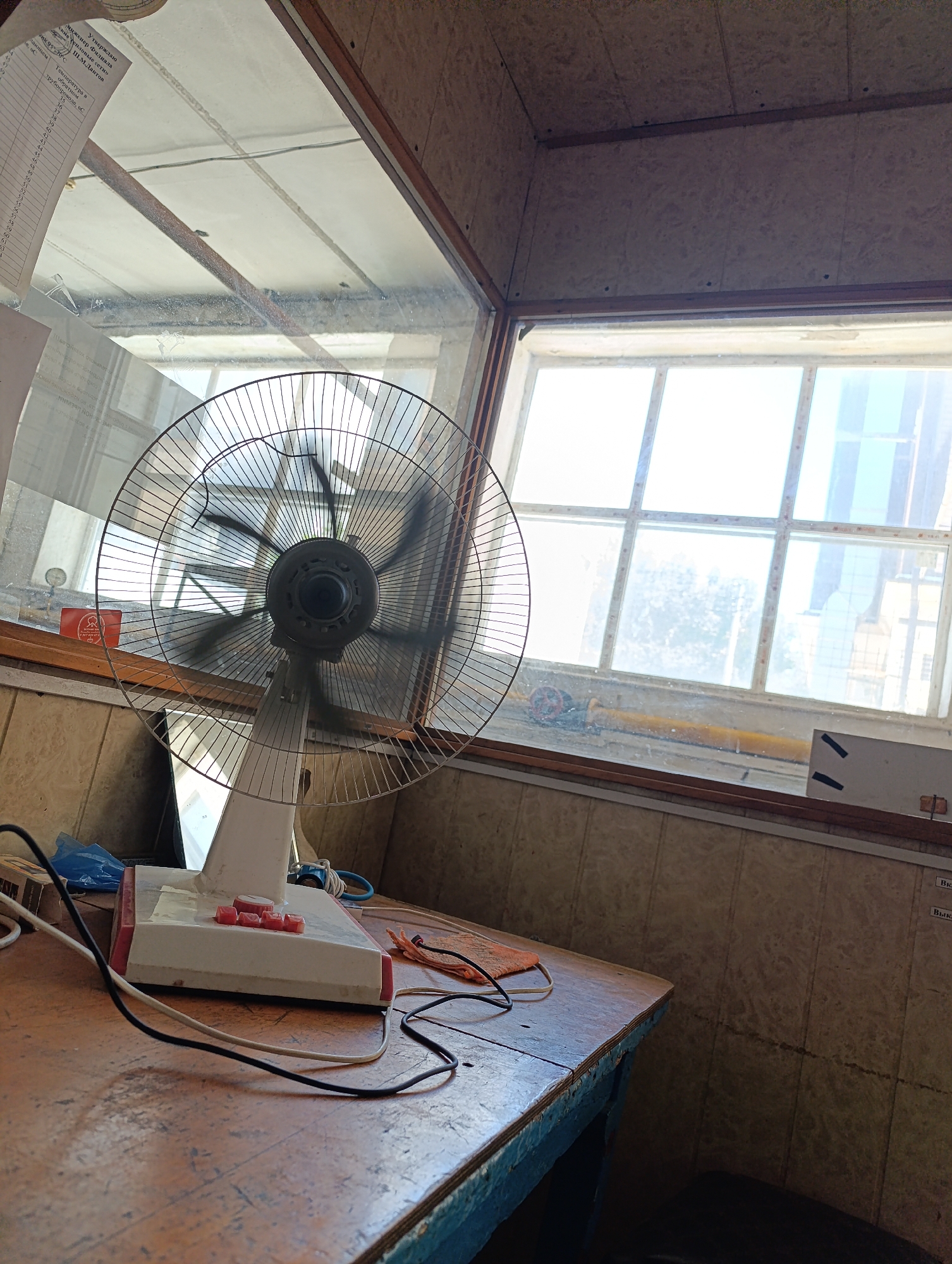 The image shows an indoor scene, likely a room with a window. A white oscillating fan with a metallic grille is positioned on a wooden table in the foreground. The fan is in operation, as indicated by the blurred blades. The table is cluttered with various items, including electrical cords, a blue plastic bag, and a small red object that may be a button or tag. The room features tiled walls and a wooden ceiling with visible beams and conduits. Through the window, a bright outdoor scene is visible, with a grid-like structure and what appears to be a building or industrial area in the distance. The overall impression is of a functional, somewhat utilitarian space. Without further information or context, it is not possible to definitively place this scene in Izberbash, Russia, or any other specific location.