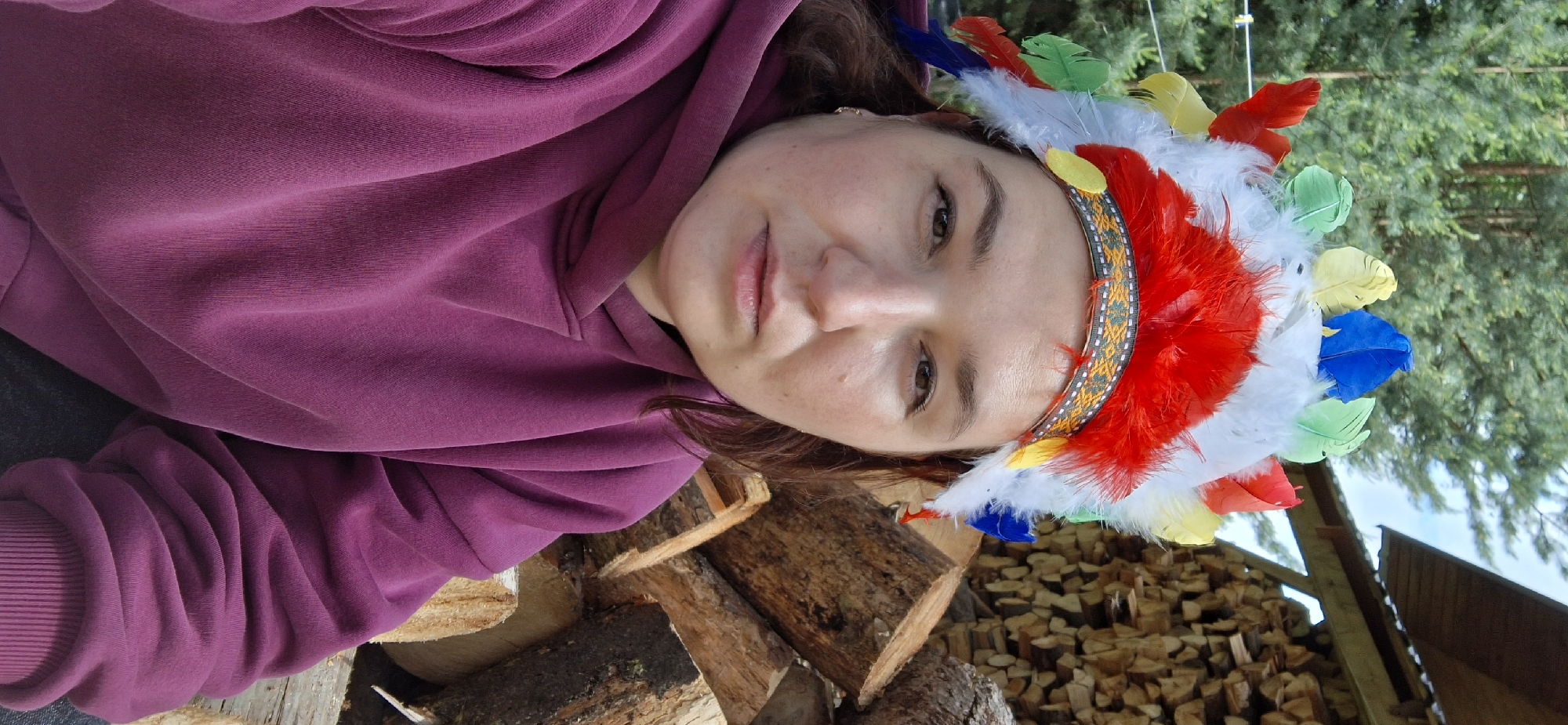 A person with light skin and brown hair is wearing a colorful feathered headdress. She is wearing a purple hooded sweatshirt and is positioned in front of a woodpile consisting of logs and chopped firewood. Background vegetation and a building are present. The city of Прикамский is not discernible in the image.