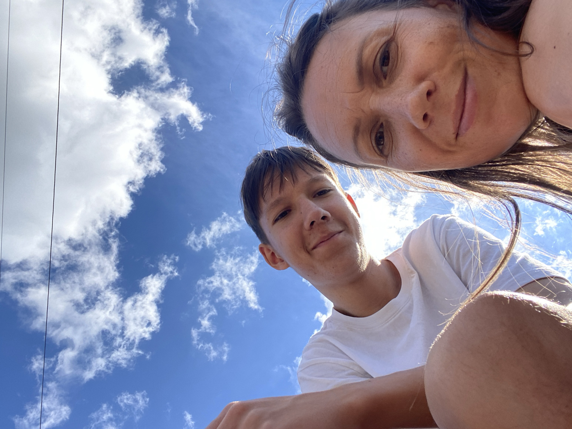 A low-angle, wide shot captures two individuals, a male and a female, against a bright blue sky dotted with white clouds. The male, on the left, wears a white t-shirt and is looking upwards and to the right with a slight smile. His left arm is visible in the foreground, bent at the elbow. The female, on the right, is closer to the camera and is looking downwards and to the left with a gentle smile. Her right shoulder and arm are visible, and her hair is being blown by the wind. The overall lighting suggests it is daytime, possibly outdoors in an urban environment given the faint power line visible across the sky. There is no indication of the city of Tashla, Russia.