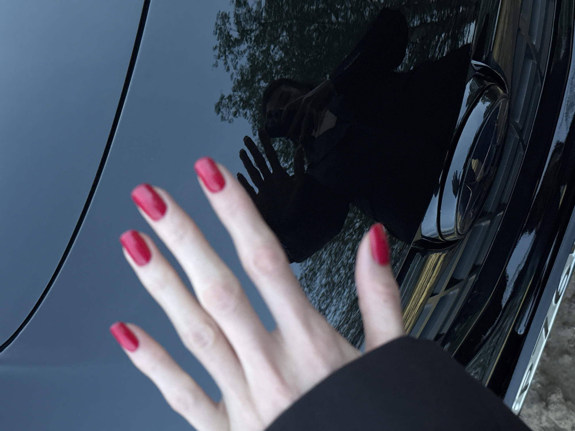 A close-up shot presents a left hand with red nail polish extended towards the glossy surface of a black car hood. The reflection on the car hood reveals a person holding a phone, possibly taking the photograph, along with the blurred image of trees overhead. The car's front grill is partially visible in the background. The location is not discernible from the image content, nor is Noviy, Russia.