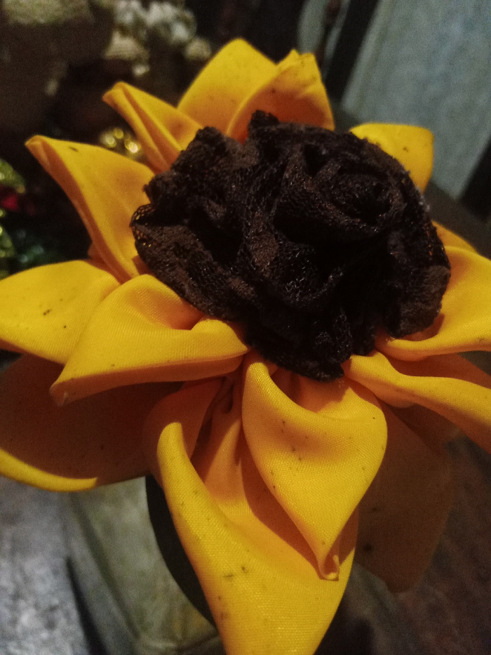 A close-up presents a handcrafted sunflower. The petals are fashioned from yellow fabric, some with noticeable dark spots. A tightly bunched, dark brown, mesh-like material forms the flower's center. The lighting is dim, casting shadows across the petals. A blurry background suggests the presence of other objects, but the sunflower dominates the view. The overall impression is one of a decorative, handmade item.