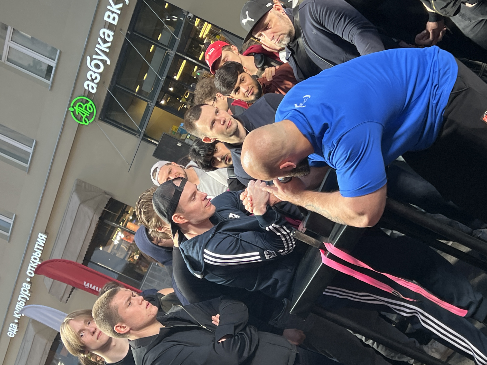 The image shows a group of people gathered outdoors around a table where two men are engaged in an arm wrestling match. The background features a building with signage in Russian, identifiable as the "Azbuka Vkusa" supermarket chain, a well-known presence in Moscow, Russia. This indicates the scene's location within Moscow. The surrounding environment appears to be an urban street. The participants and spectators are dressed in casual clothing, with some wearing Adidas apparel. The atmosphere suggests a local event or contest.