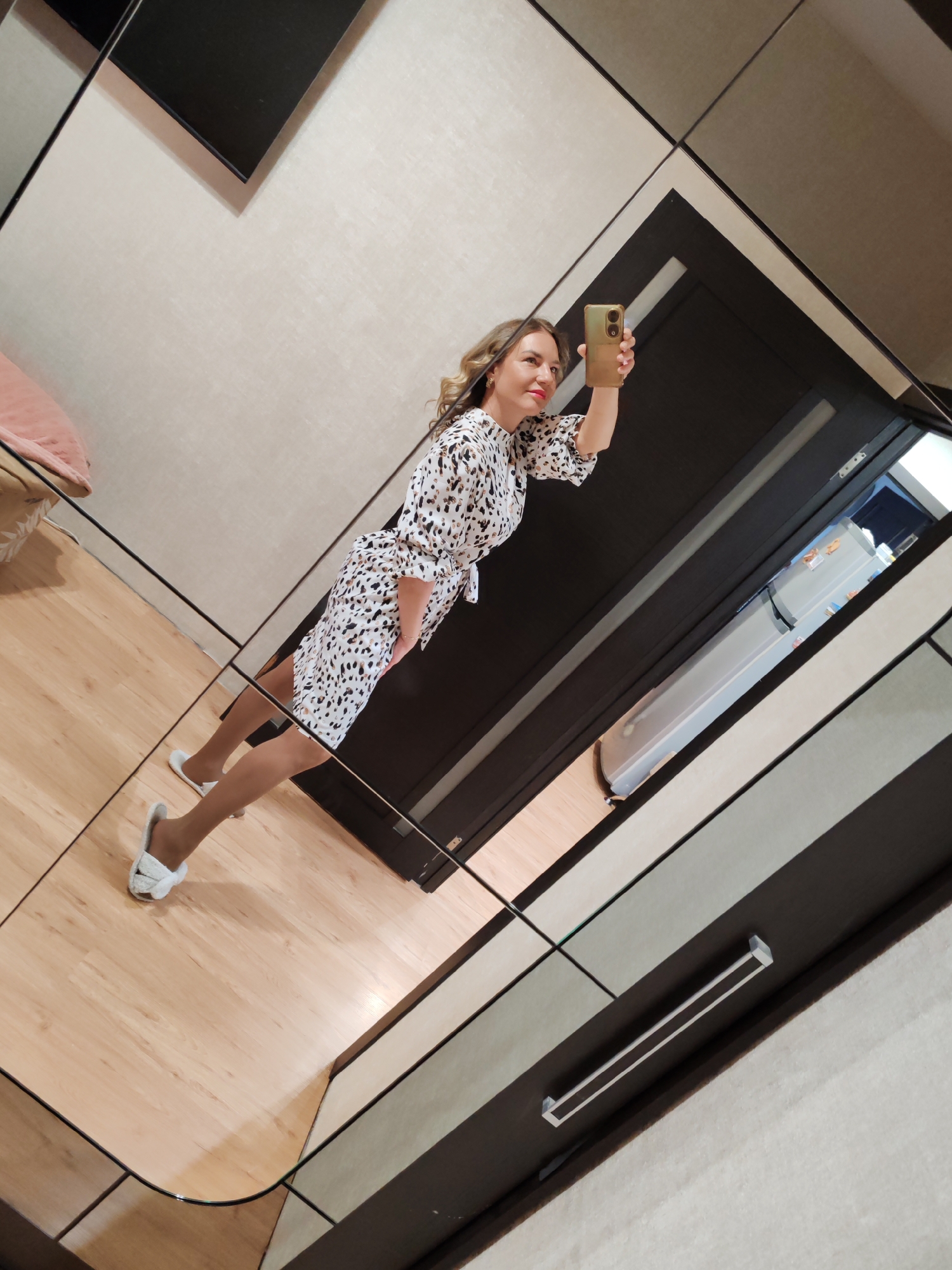 A woman is taking a selfie in a room. She is wearing a patterned dress, pantyhose, and white slippers. She holds a phone in her right hand and smiles towards the camera. The room appears to be a bathroom or dressing area with a mirrored wall reflecting a doorway and a glimpse of a refrigerator. The flooring is light wood. The room's decor features dark and light patterned panels on the walls. There is a pink object in the left corner of the frame. The location is possibly Petropavlovsk-Kamchatsky, Russia.