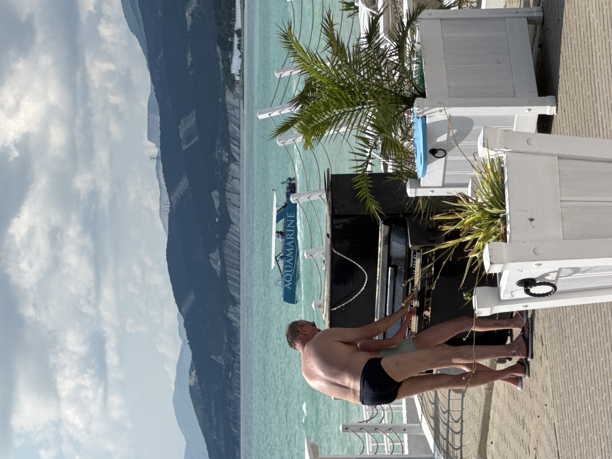 In Novorossiysk, Russia, on a sunny day, a man and a child stand at an upright piano on a paved walkway near the sea. The man, wearing black swim trunks, is positioned behind the child, who is wearing a light-colored garment. Both appear to be interacting with the piano keys. In the background, the turquoise sea is visible, with a boat labeled "AQUAMARINE" cruising on the water. Mountains form a scenic backdrop under a partly cloudy sky. White planters with greenery and a white railing with ropes are in the foreground, suggesting a waterfront promenade or resort area.