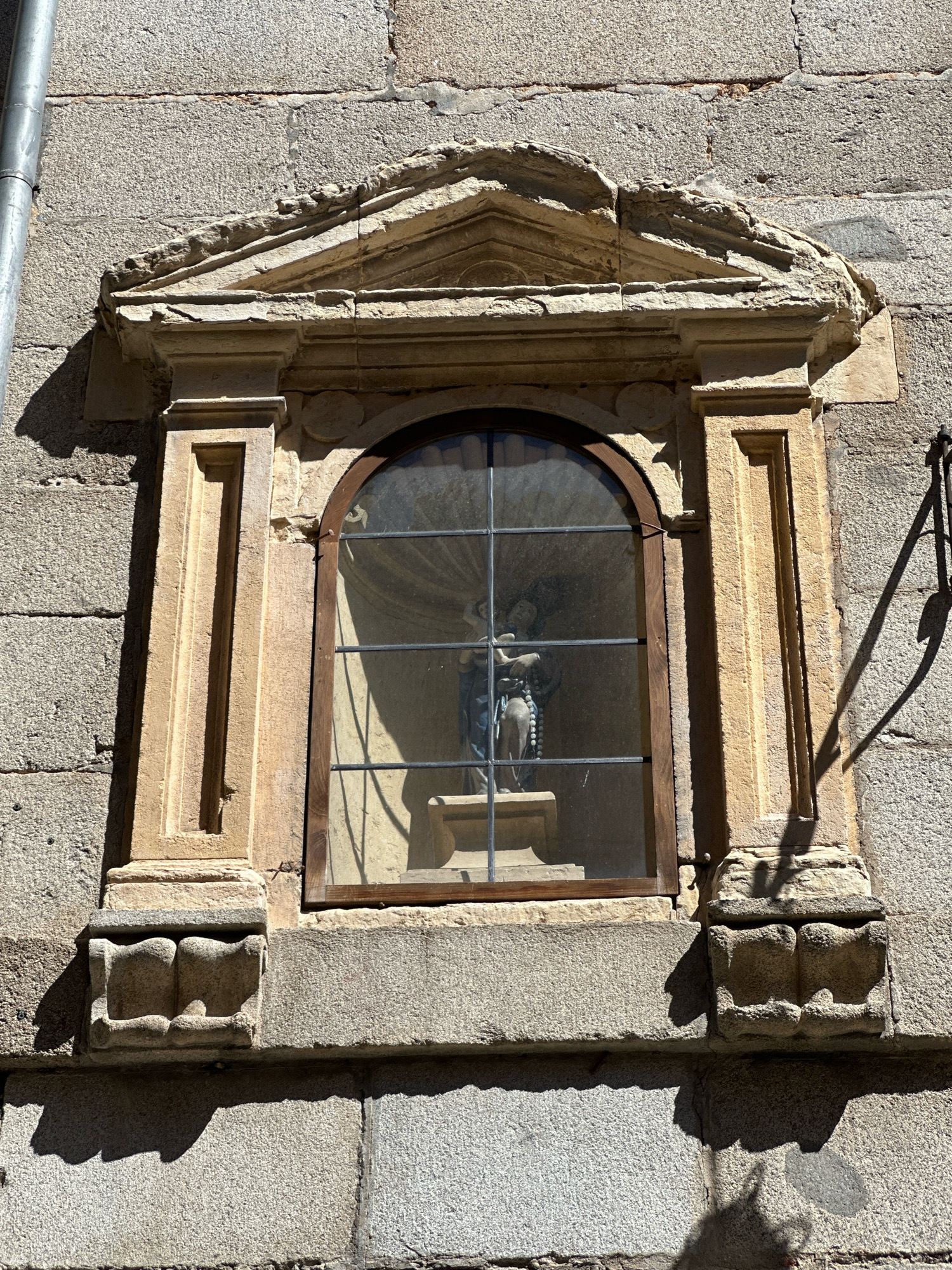 The image shows an ornate stone niche built into a stone wall. The niche contains a statue of a religious figure, possibly the Virgin Mary, holding a rosary. The statue is enclosed by a glass pane with metal bars. The niche is decorated with carved architectural elements, including a pediment at the top and pilasters on the sides. The stone wall is rough-hewn and appears weathered. The scene is lit by sunlight, casting shadows on the wall and niche. This niche is located in the municipality of La Lastrilla, Spain, which is part of the province of Segovia.