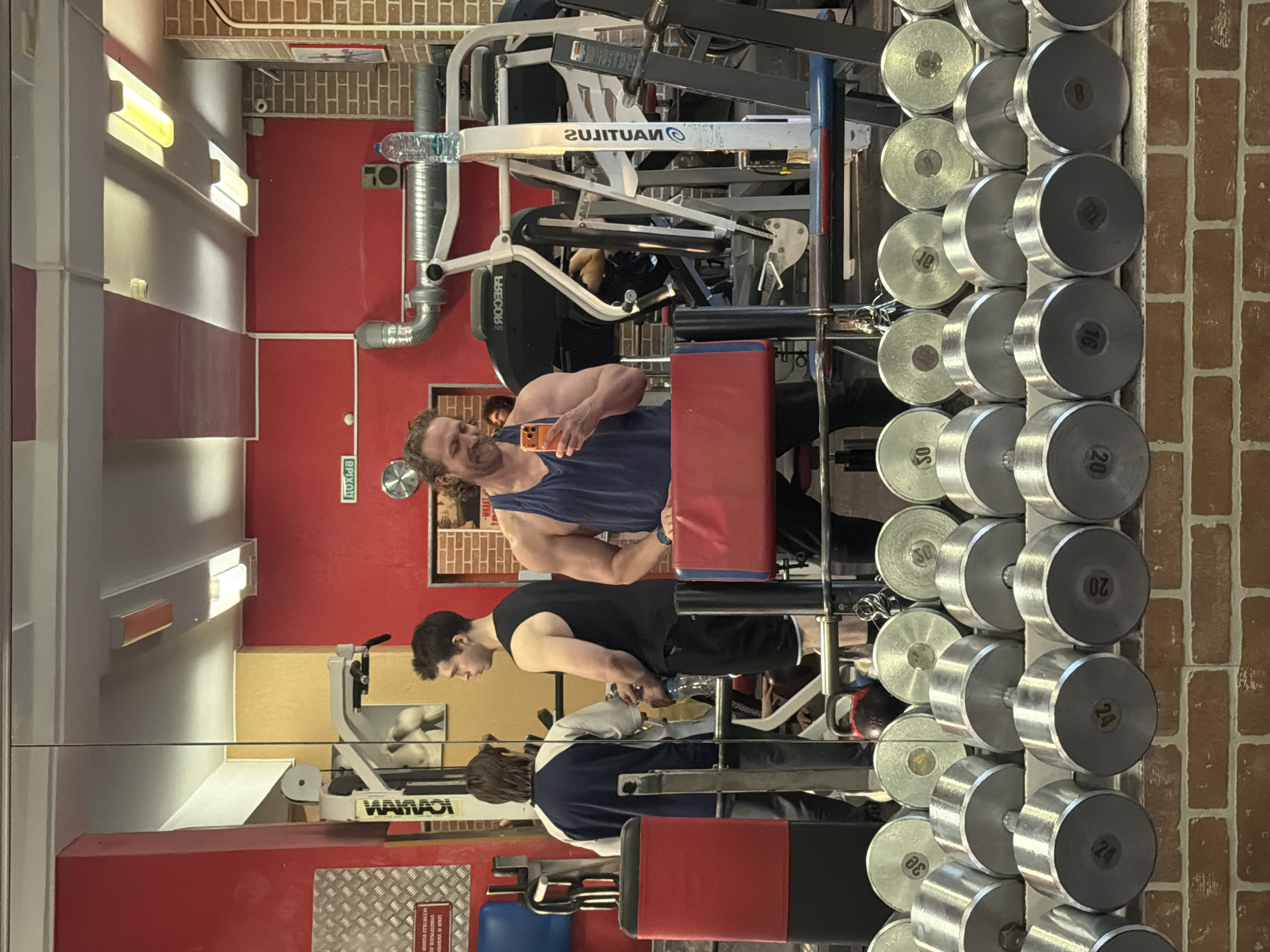 The image depicts the interior of a gym, likely in Saint Petersburg, Russia based on the Cyrillic text "ВЫХОД/ВРІХОД" on an exit sign, as seen in the reflection of a mirror. Three men are visible. One man in a blue tank top is holding a phone and appears to be taking a selfie. Another man is wearing a black tank top. The third man is wearing a blue and grey sweatshirt. They are near various pieces of gym equipment, including a Nautilus machine, a Ваймади bench press, and a dumbbell rack loaded with various weights. The dumbbells range from 16kg to 30kg. The gym features a red accent wall, a brick wall, and overhead fluorescent lighting. A water bottle is on a Nautilus machine. The scene suggests an active environment with people working out and potentially documenting their gym session.