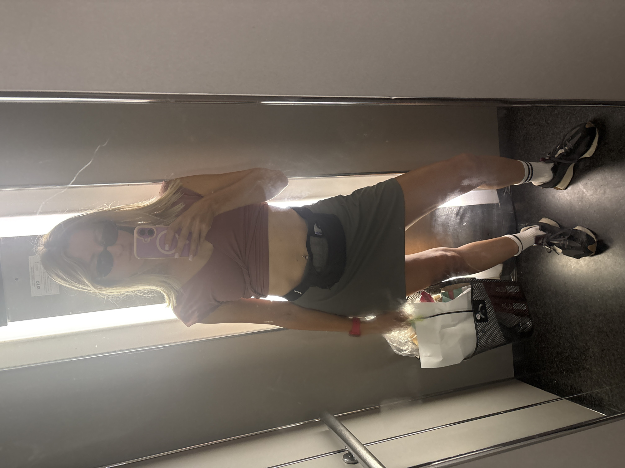 A woman takes a mirror selfie in what appears to be a locker room or dressing room, possibly at a train station in Valencia, Spain. She is wearing a mauve crop top, a dark grey skirt, a black fanny pack, white socks with blue stripes, and dark sneakers. She has sunglasses on and is holding a purple-cased phone to take the photo. Her hair is blonde and falls to her shoulders. To her left, a black mesh basket filled with items, including white paper bags, rests on the floor. The environment is utilitarian with grey walls and a dark floor.