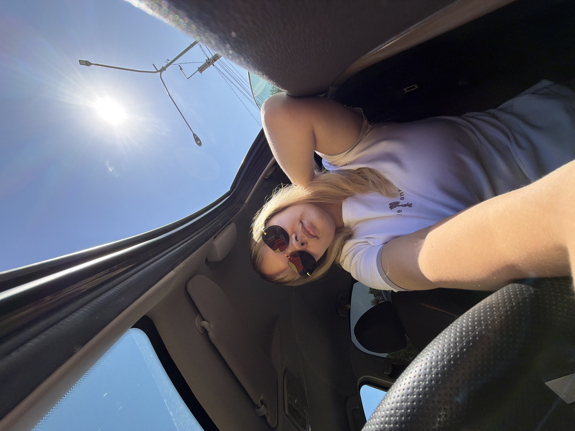 A low-angle selfie photograph, taken from inside a vehicle, shows a woman wearing sunglasses looking down at the camera. The sky is visible through the car's open sunroof, and the sun is shining brightly, partially obscured by a streetlight. The woman is wearing a white T-shirt. The interior of the car is visible, including the dashboard, rearview mirror, and front seats. The location is likely outdoors, and the bright sunlight suggests daytime. The city of Chaikovsky, Russia, is not confirmed.