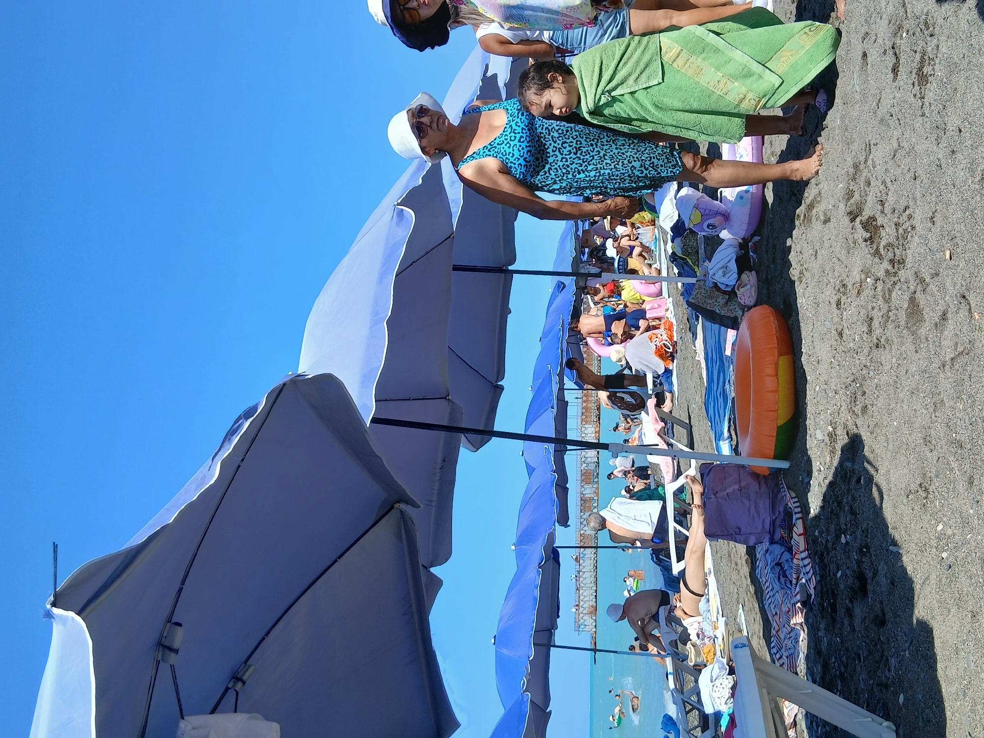 A sunny day at the beach in Zharbulak, Kazakhstan, is depicted. The foreground shows dark, pebbly sand with several beach umbrellas providing shade. In the mid-ground, a group of people are visible under the umbrellas, some sitting on lounge chairs, others standing. A woman in a blue leopard-print swimsuit and a white hat is standing with a young child wrapped in a green towel. Behind them, more people are relaxing on the beach and in the shallow water. The sea is calm and blue, with a long pier visible in the distance. The sky is clear and bright blue. Several inflatable toys, including a pink dolphin and orange and green rings, are scattered on the sand.