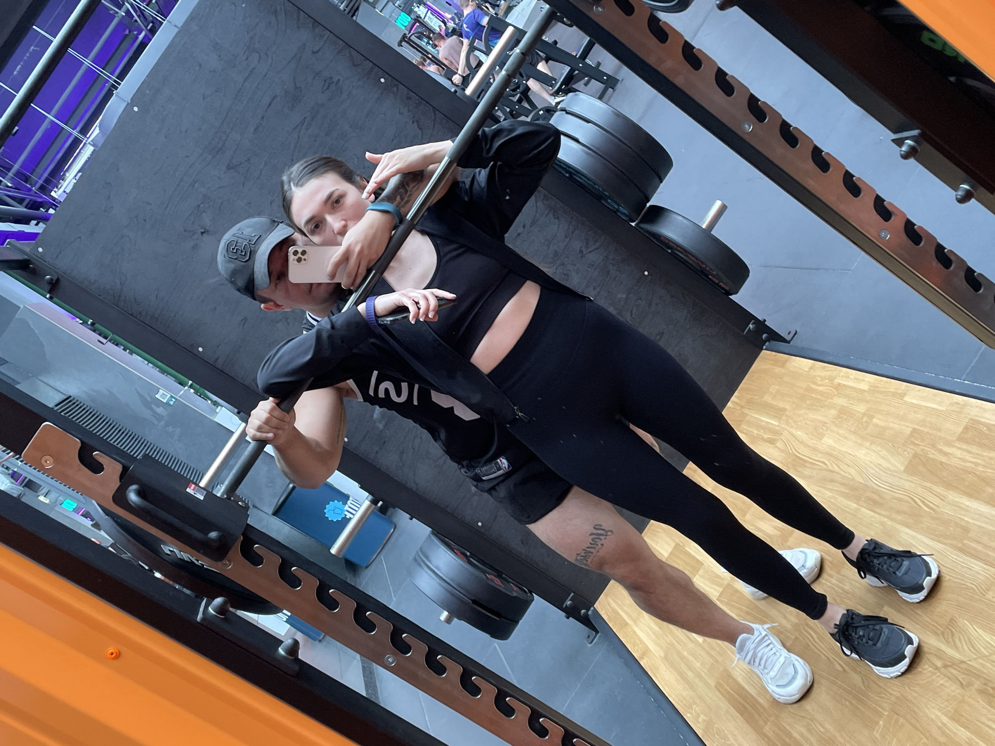 The image shows a man and a woman taking a selfie in a gym in Kazan, Russia. The gym is equipped with weightlifting equipment, including a squat rack with weight plates loaded on it. The man is wearing a baseball cap, a t-shirt, and shorts. The woman is wearing athletic attire, including a sports bra and leggings. They are both holding the camera phone to take the selfie. The floor is light brown wood. There are people working out in the background.