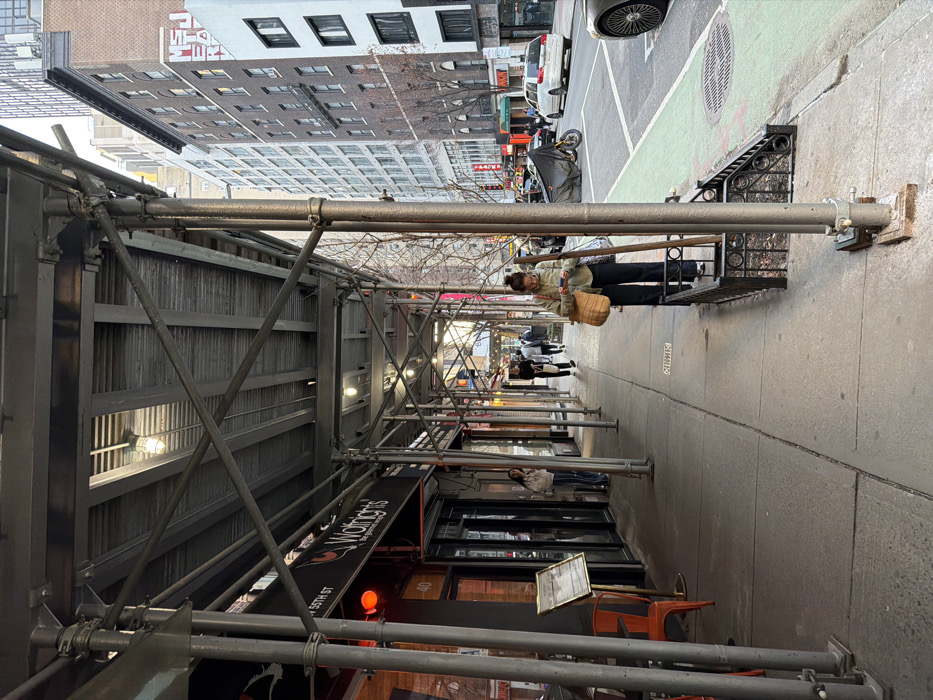 Location: West 55th Street, New York City, United States of America.
Scene: A street-level view of a New York City sidewalk covered by a dark grey metal scaffolding structure. The sidewalk runs parallel to a street featuring vehicle traffic and a green-painted bicycle lane. Buildings of various heights and architectural styles line both sides of the street under an overcast sky.
Details: The scaffolding's corrugated metal roof is supported by numerous metal poles and cross-braces, with lights illuminating the walkway beneath. On the left, a storefront for "Wolfrigh's" restaurant, marked with "W 55TH ST" and address "40", features a dark facade, red accents, glass doors, an outdoor menu stand, and an orange chair. A female, with long dark hair, wearing a white top and striped pants, stands near the restaurant entrance, looking right. Further down the sidewalk, two male individuals walk away from the camera. In the foreground right, a female with pulled-back brown hair, wearing a light olive-green jacket, dark pants, and white sneakers, stands by a low decorative metal railing, holding and looking at a smartphone; a woven straw bag hangs from her left shoulder. The street contains several vehicles: a partially visible dark luxury car in the immediate foreground right, a black tarp-covered motorcycle or scooter parked near the green bike lane, and a silver minivan in the middle distance. Bare trees are visible along the street. A multi-story brick building across the street displays partial red text indicating "METROPOLITAN OPERA".