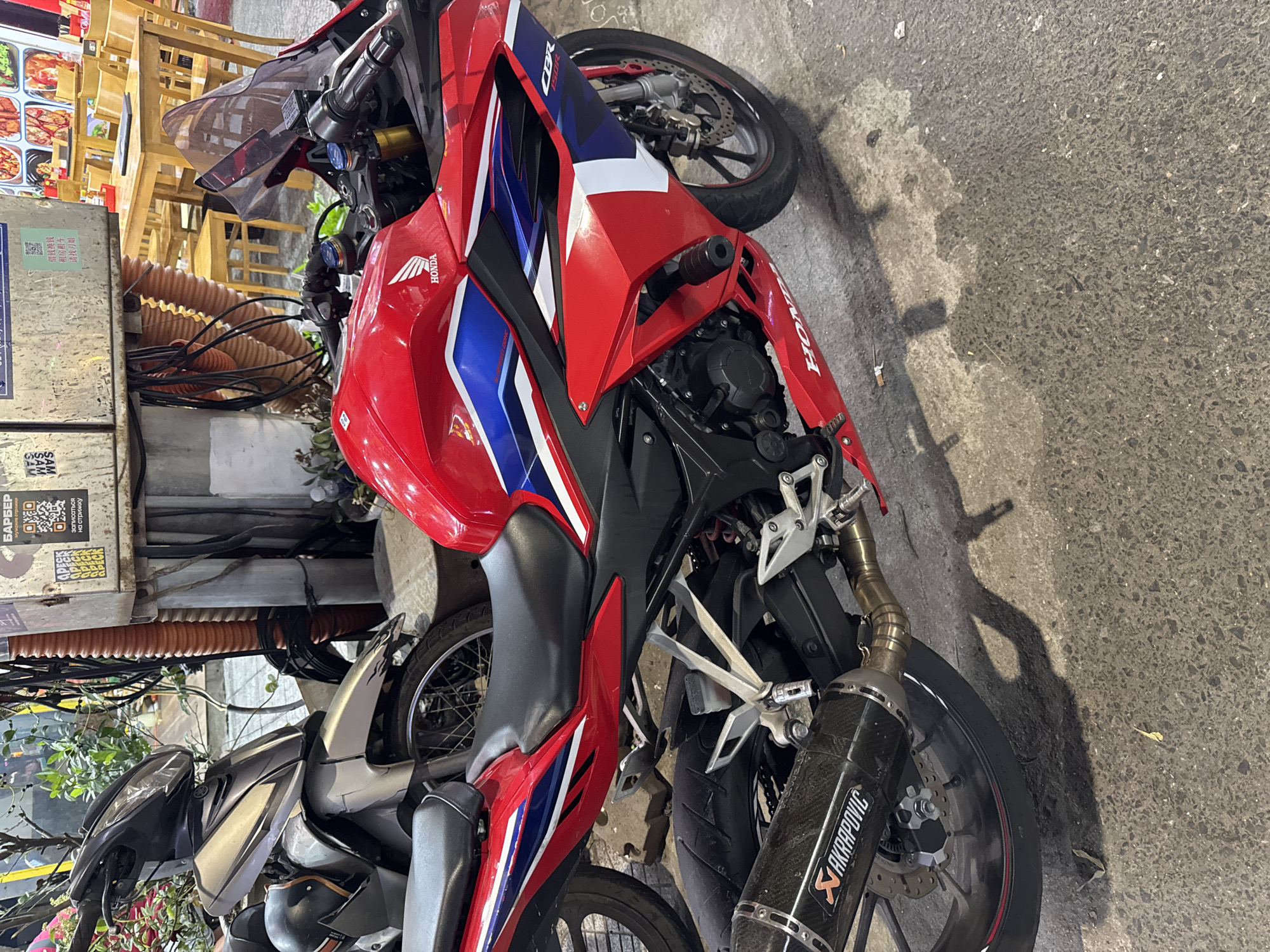 The image shows a Honda CBR150R motorcycle parked on a city street. The motorcycle is red, white, and blue. It has an Akrapovic exhaust. Parked to the left of the Honda is another motorcycle. A transformer is next to the bikes, with signs affixed advertising a barber and services offered by someone named "刀姐." The location is likely in the city of Đà Nẵng, Vietnam, based on the presence of Vietnamese text on the signs.