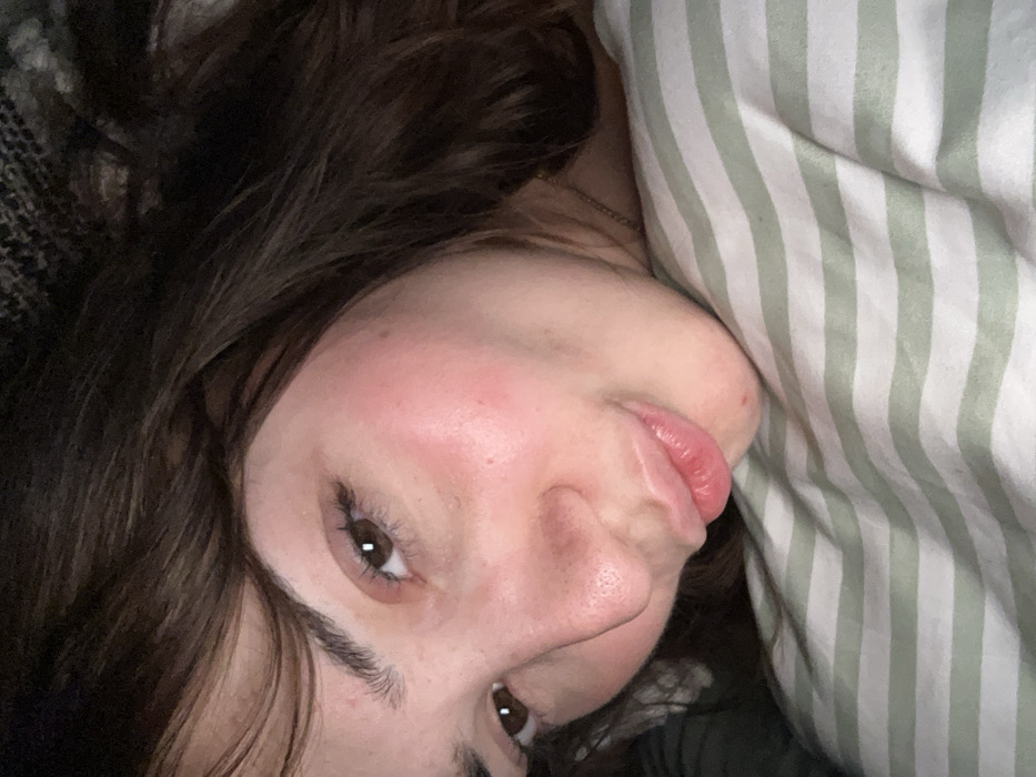 A woman with brown hair and fair skin is lying on a bed. Her head rests on a pillow with green and white stripes. Her eyes are open, and her lips are slightly parted. The camera angle is from above, capturing her face and upper body. Part of a dark, patterned blanket is visible in the background.