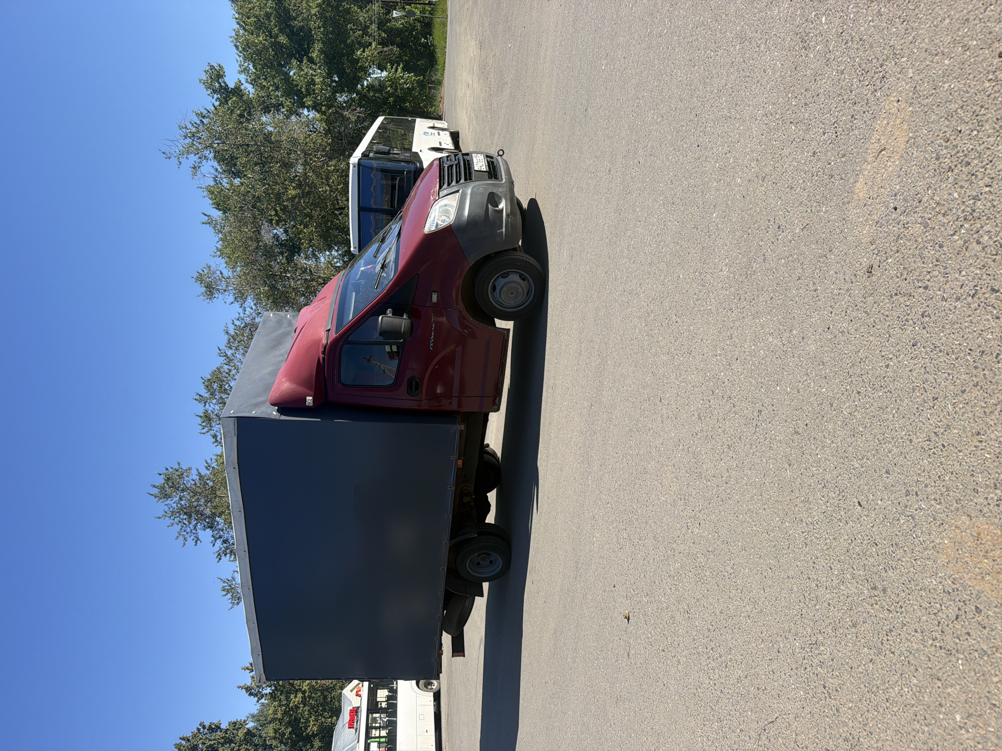 A red truck with a dark gray cargo box is parked on an asphalt lot under a clear blue sky. The truck is facing right and is positioned in the foreground. Behind the truck, two white buses are parked side-by-side. Trees line the background of the scene. The location appears to be in the city of Chaikovsky, Russia, possibly a bus depot or parking lot. No people are visible in the image.