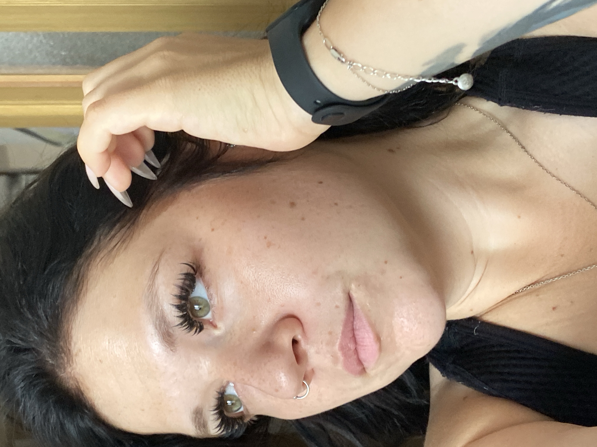 A close-up, chest-up, medium shot captures a young woman with dark hair and light green eyes, looking off to the left. Her face is lightly powdered, with freckles visible on her cheeks and nose. She has pronounced false eyelashes, a silver hoop nose ring, and is wearing a black tank top. Her right hand is raised, with long, light-colored acrylic nails resting near her temple and against her hair. A black smartwatch is on her left wrist, accompanied by a delicate silver chain bracelet. A thin silver necklace with a small pendant is visible against her neck. The background is out of focus, showing a blurred vertical golden structure to the right, suggesting an indoor setting, possibly a home. The image does not provide any specific information about the city of Unknown, Russia.