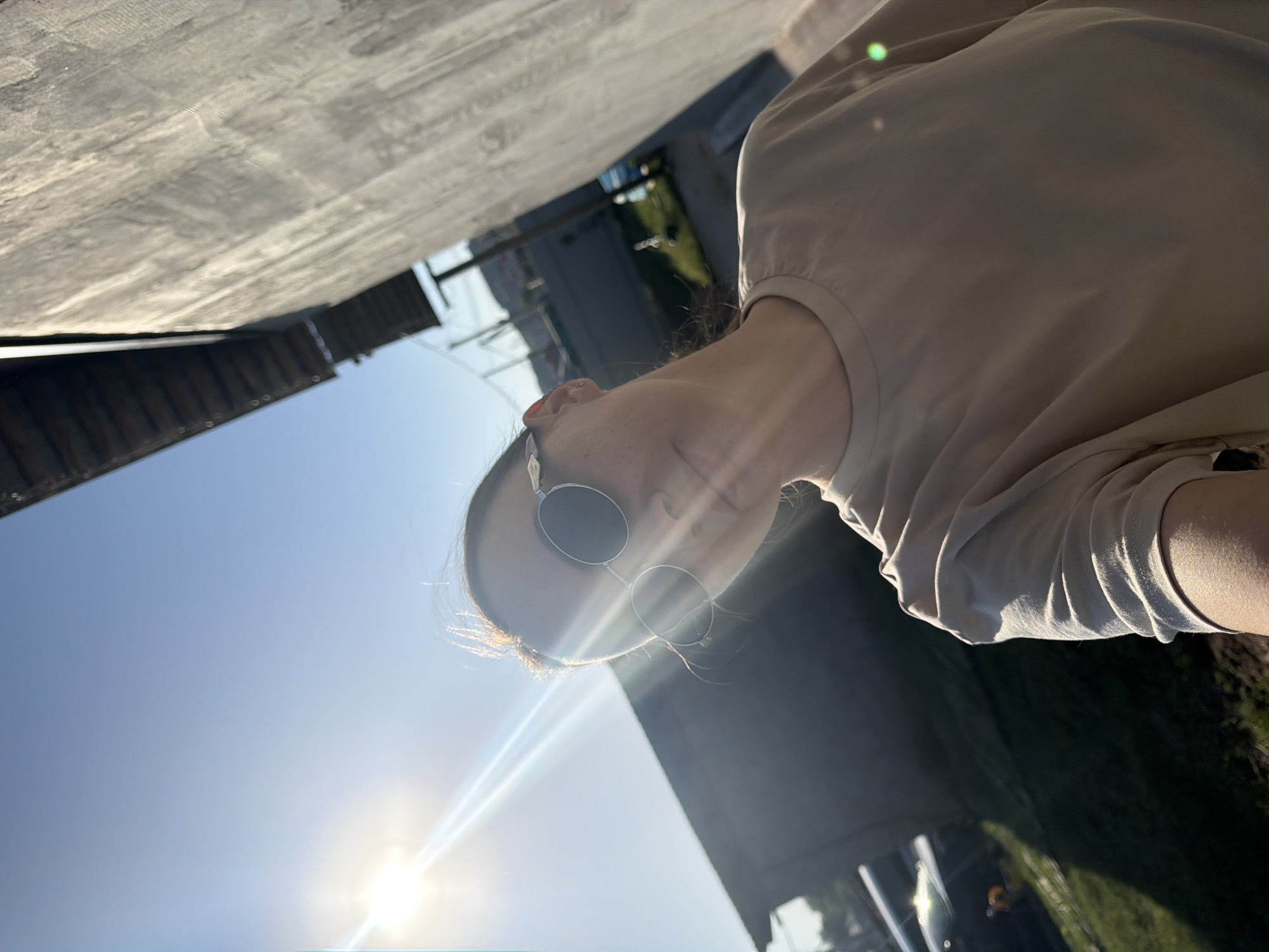 A woman with sunglasses is outdoors on a sunny day. She is wearing a light-colored shirt. The location appears to be in an urban environment.