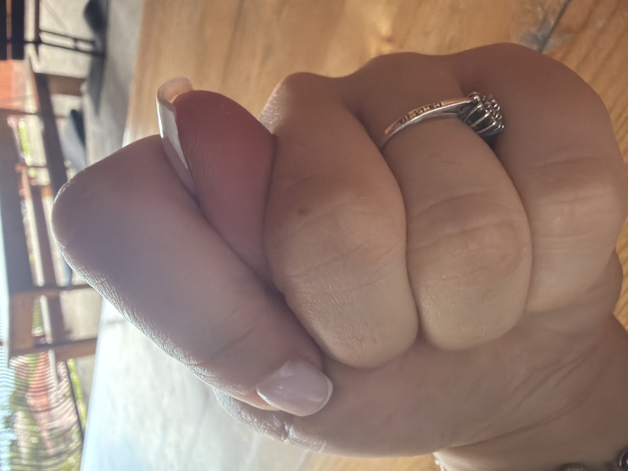 A close-up, high-angle shot shows a person's right hand clenched in a fist, with the knuckles pressing into the palm. The fingers are covered in light pink nail polish. A silver ring with a cluster of small stones is visible on the ring finger. The background consists of a wooden table and blurred outdoor scenery with tables and chairs. The overall lighting suggests an indoor or shaded outdoor setting.