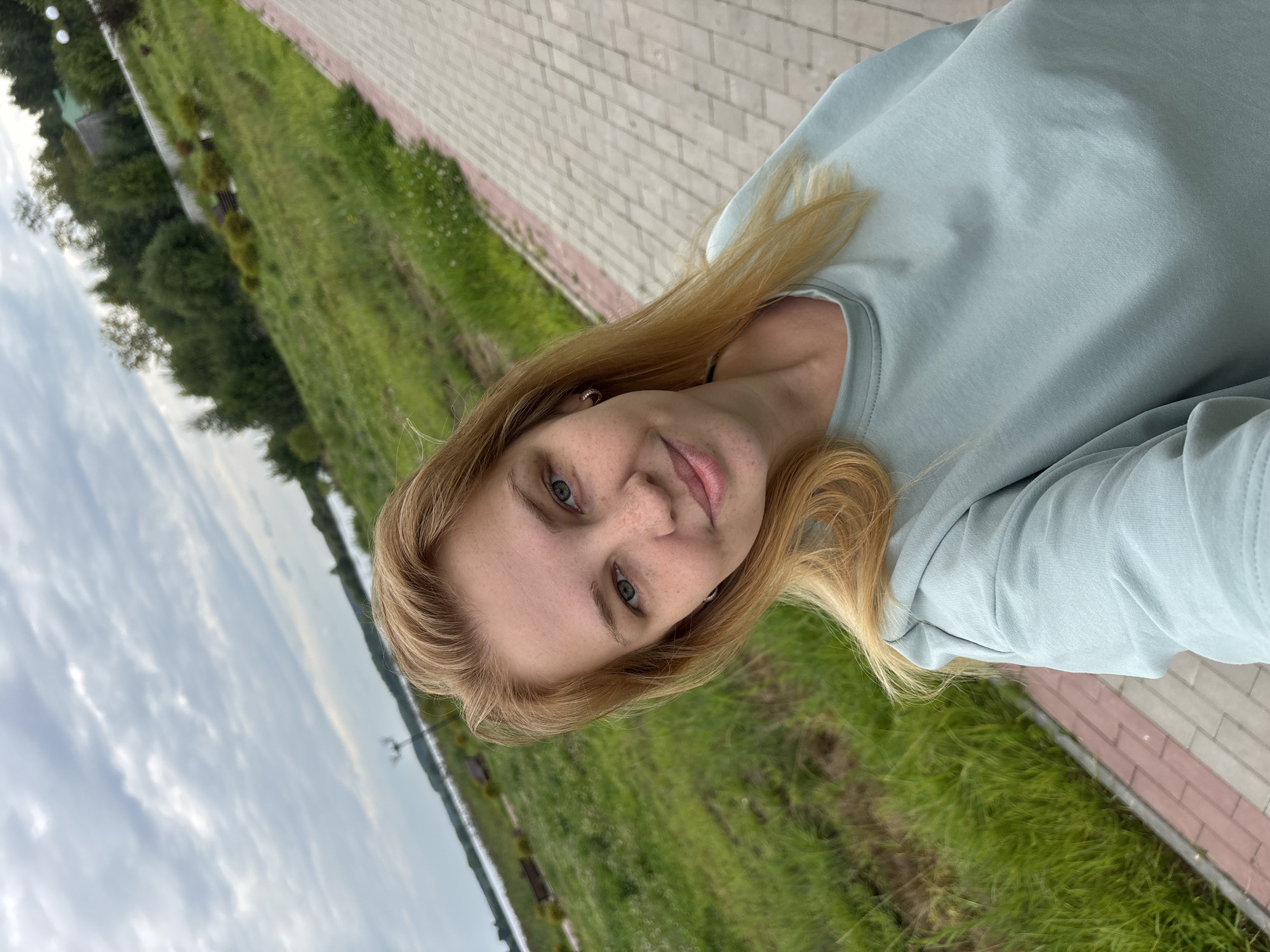 A young woman with blonde hair and freckles is taking a selfie outdoors. She is wearing a light blue sweatshirt. The background shows a grassy area, a paved path, a body of water, and trees. The sky is cloudy. The image appears to be taken in Breytovo, Russia, as indicated by the presence of a lamppost with a Cyrillic sign and the general landscape which is consistent with the region.