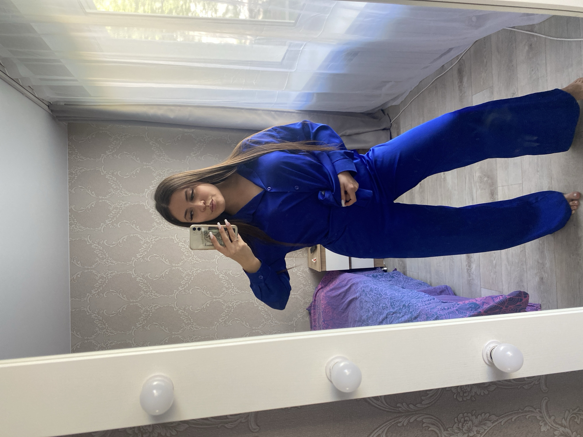 A young woman is taking a selfie in a mirror. She is wearing a blue pajama set. She is holding a phone with a clear case. The background includes wallpaper with a floral pattern, white curtains, a window, and a bed with a patterned purple and blue cover. The location appears to be an interior space, possibly a bedroom. There is no verifiable evidence to confirm if this photograph was taken in the city of Chaikovsky, Russia.