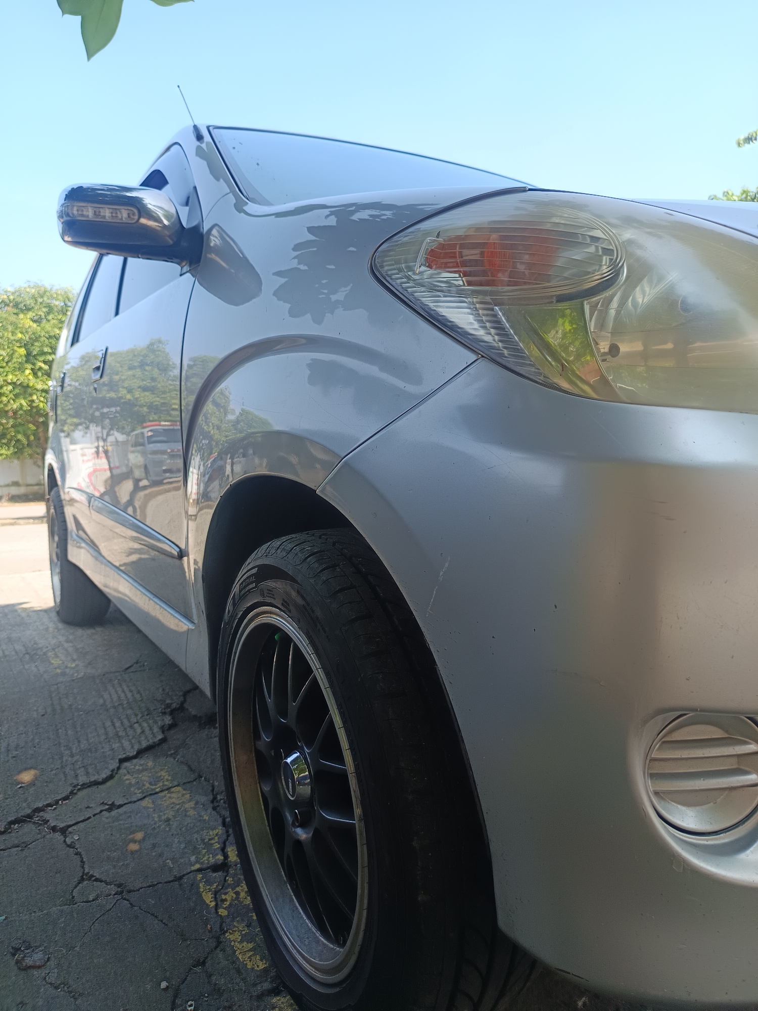 A low-angle, close-up shot of the front passenger side of a silver, four-door hatchback car, likely parked on a cracked asphalt surface with faded yellow lines. The car's headlight and side mirror are visible, along with part of the front fender and wheel. The sky above is clear and blue. The scene appears to be outdoors in a residential or commercial area in Karangtengah, Indonesia, possibly a driveway or parking lot, indicated by the cracked asphalt and the reflection of a building and other cars in the car's door. The overall impression is that of a sunny day.