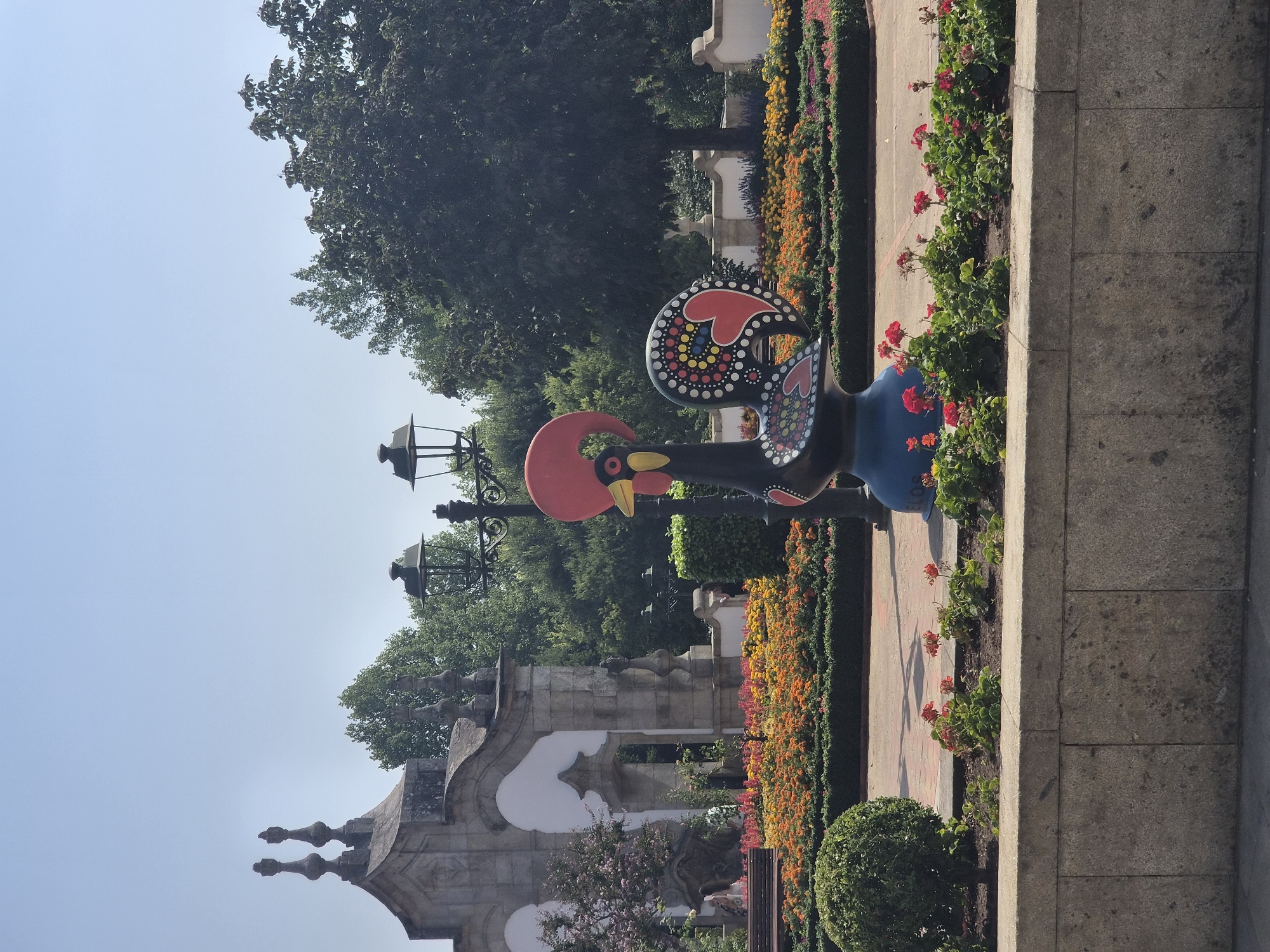The image depicts a colorful statue of a rooster, a prominent symbol of Barcelos, Portugal, situated in a public garden. The rooster statue, predominantly black with red, yellow, blue, and white decorative patterns, stands on a blue base in the foreground. Behind the statue, a neatly manicured garden features rows of vibrant orange and yellow flowers bordered by green hedges. To the left, an ornate stone archway, characteristic of Portuguese architecture, stands adorned with sculpted finials. Several lampposts are visible, adding to the park-like ambiance. The background is filled with lush green trees under a clear blue sky, suggesting a bright, sunny day. The garden setting implies a public space, likely a tourist attraction or a park within the city of Barcelos, a city renowned for its pottery and folk art, including the iconic Rooster of Barcelos. There are no people clearly visible interacting with the statue or the environment. The overall scene is one of a well-maintained outdoor public space showcasing local cultural iconography.