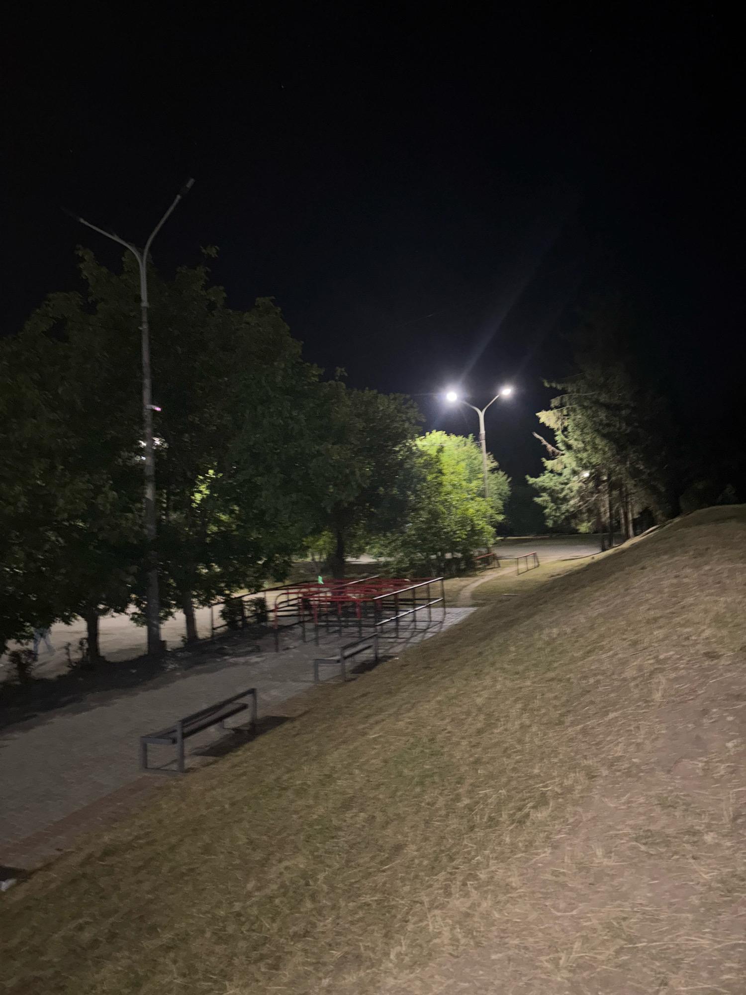 The image depicts a park scene at night in Ternopil, Ukraine. Two streetlights illuminate a pathway and a grassy slope. Several trees are visible, casting shadows in the artificial light. In the foreground, a park bench sits on the paved pathway, which runs alongside the grassy incline. Further back, red metal railings and structures, possibly part of playground equipment or seating, are visible. The overall impression is of an empty public space after dark, lit by artificial lighting.
