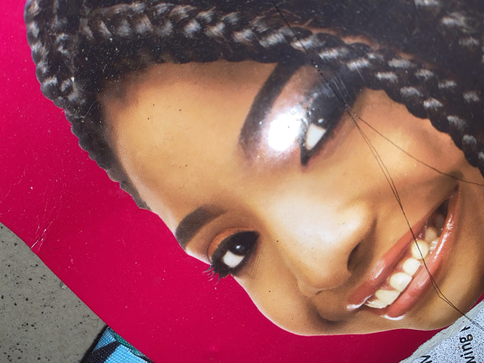 The image shows a close-up of a person's face, with a focus on their eye and mouth. The person has dark skin and is wearing makeup, including eyeshadow and lipstick. Their hair is styled in braids. The background is a vibrant pink color, with some texture and a hint of a gray, speckled surface on the left side. There is also a torn piece of paper with some writing on it visible in the lower left corner. The overall impression is that this is a product image or a portrait. The location in Jauro Yinu, Nigeria is not discernible from the provided image.