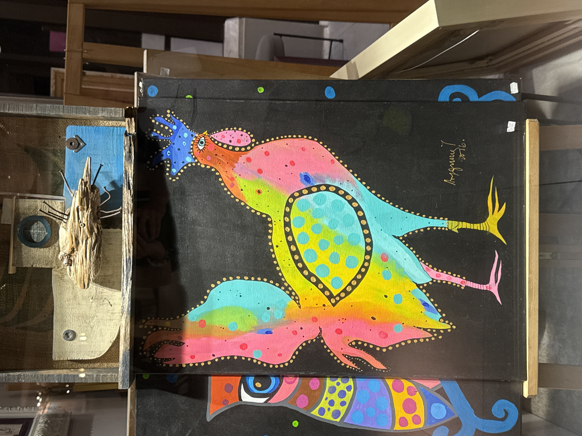 The image displays a vibrant, colorful painting of a rooster on a black background, positioned on a wooden easel. The rooster is depicted in a stylized, folk-art manner, with a rainbow gradient of colors on its body, accented with gold dots and teal and pink polka dots on its wings and tail. It has a blue, crown-like crest and large, expressive eyes. To the left of the rooster, another painting of what appears to be a stylized owl or bird is partially visible, also featuring bold colors and patterns. Above the paintings, a mixed-media assemblage is mounted, incorporating wood, fabric, and hardware, resembling a whimsical birdhouse or shelter. The overall scene suggests an art display or gallery setting. The signature on the rooster painting reads "Aygun" and "2016." The backdrop of the scene indicates an indoor environment, possibly a shop or exhibition space, with wooden frames and display structures visible. The location is identified as Budva, Montenegro.