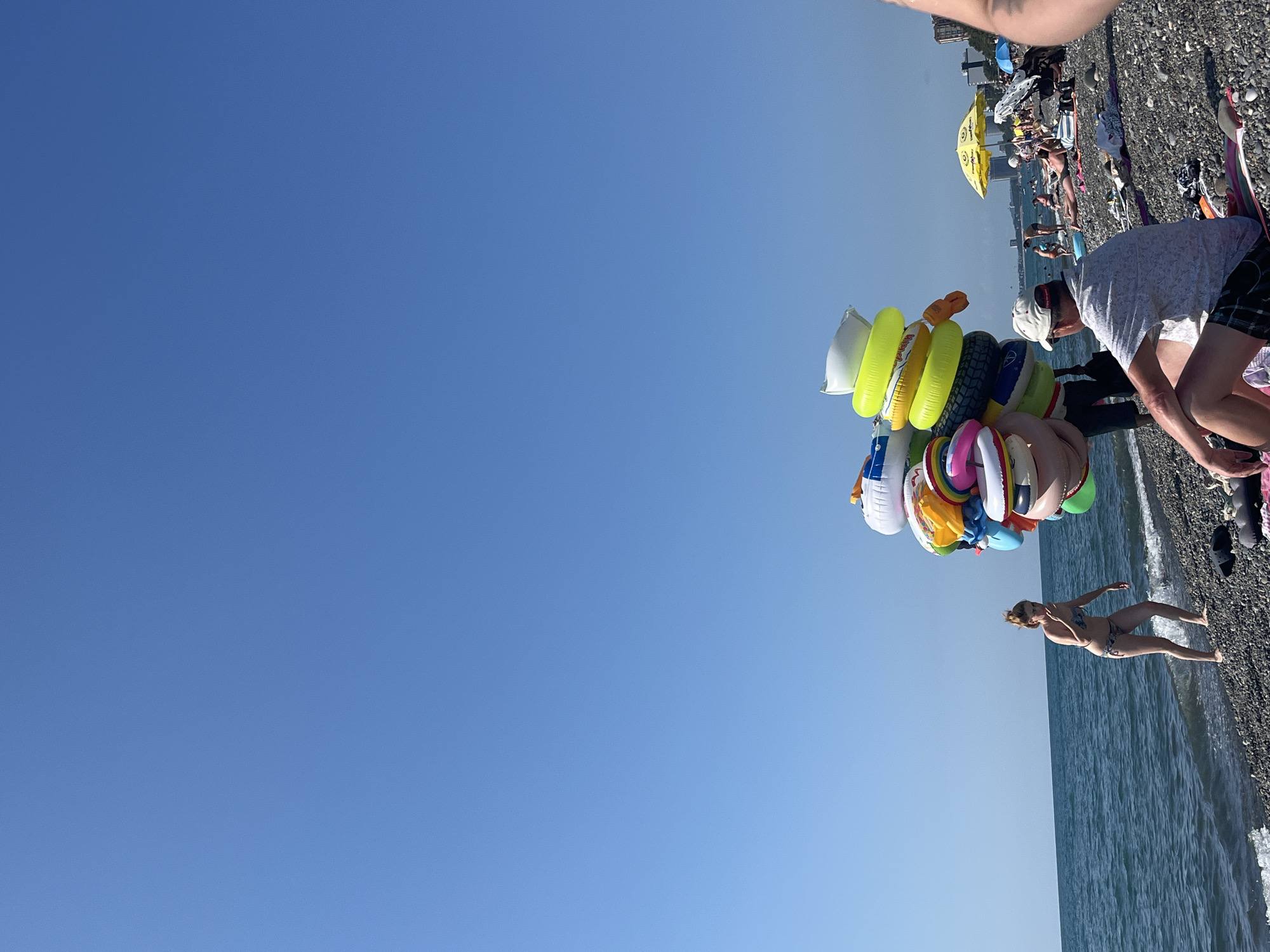 A beach scene in Kobuleti, Georgia, depicts a vendor selling inflatable rings. A man, possibly the vendor, is seen from the chest down, wearing a patterned short-sleeved shirt and a white cap. He is holding a large bunch of colorful inflatable rings. To the right, a woman in a bikini is walking on the pebbly beach near the water. The sea is visible with gentle waves. In the background, other beachgoers are seen relaxing under umbrellas, and buildings line the coast. The sky is a clear, bright blue, suggesting a sunny day, likely in the afternoon due to the strong sunlight. The overall mood appears to be relaxed and leisurely, typical of a summer day at the beach. No visible text is present in the image.