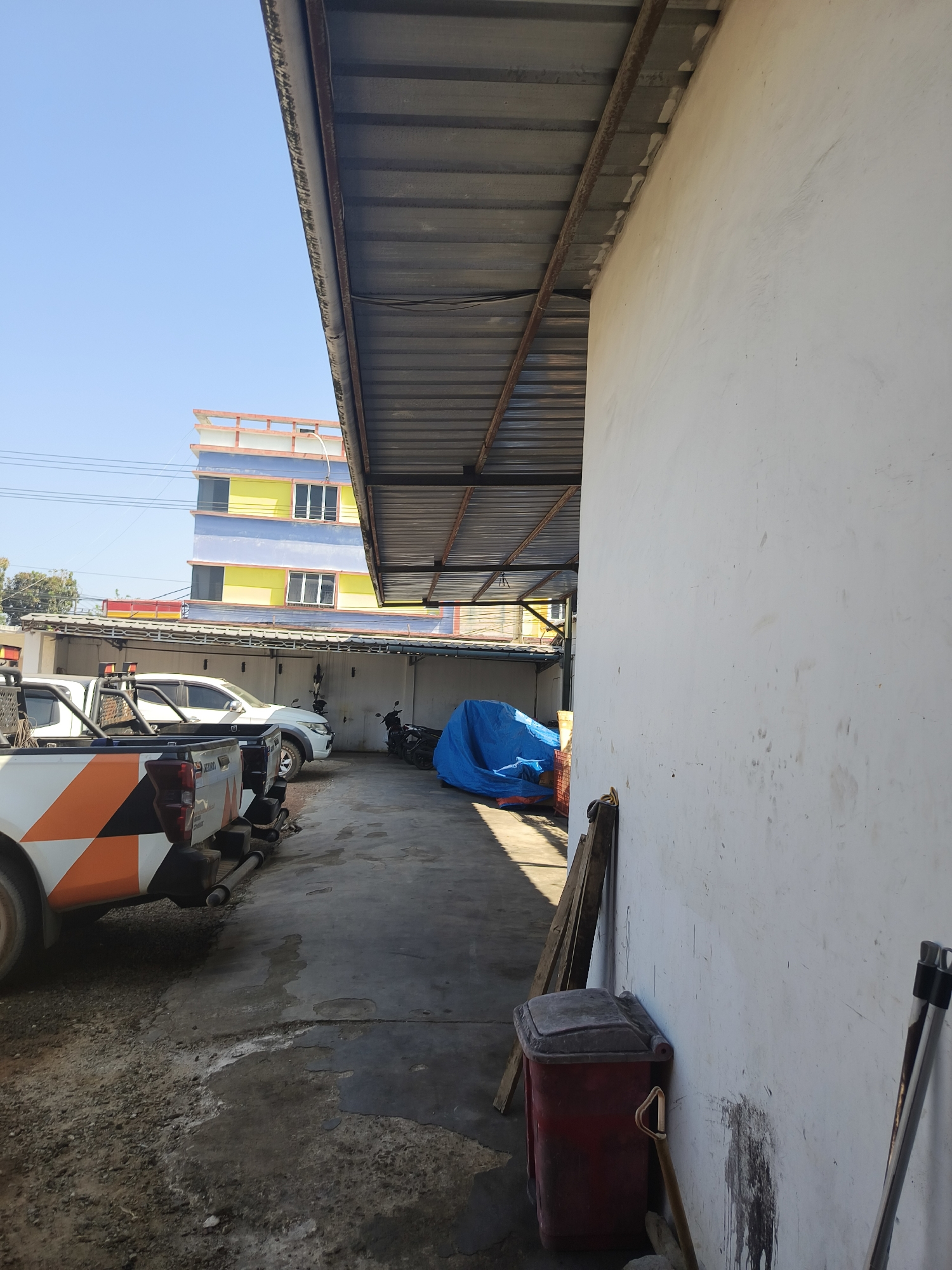 This image depicts an outdoor area, likely a storage or service space, with a white building wall on the right and a corrugated metal roof supported by metal beams extending from the left. Under the roof, several vehicles are parked. In the foreground, the rear of a white pickup truck with orange and black accents is visible. Behind it, another white pickup truck and a white SUV are parked. Further down, a blue tarp covers an object, and several motorcycles are parked. In the background, a multi-story building with yellow, blue, and orange sections is visible against a clear blue sky. The ground appears to be a mix of concrete and gravel, with some areas showing signs of dampness. A red trash bin and some wooden planks are leaning against the white wall on the right. The scene suggests a utilitarian outdoor space, possibly within an industrial or commercial area of Pangkalpinang, Indonesia. There are no people visible in the image.